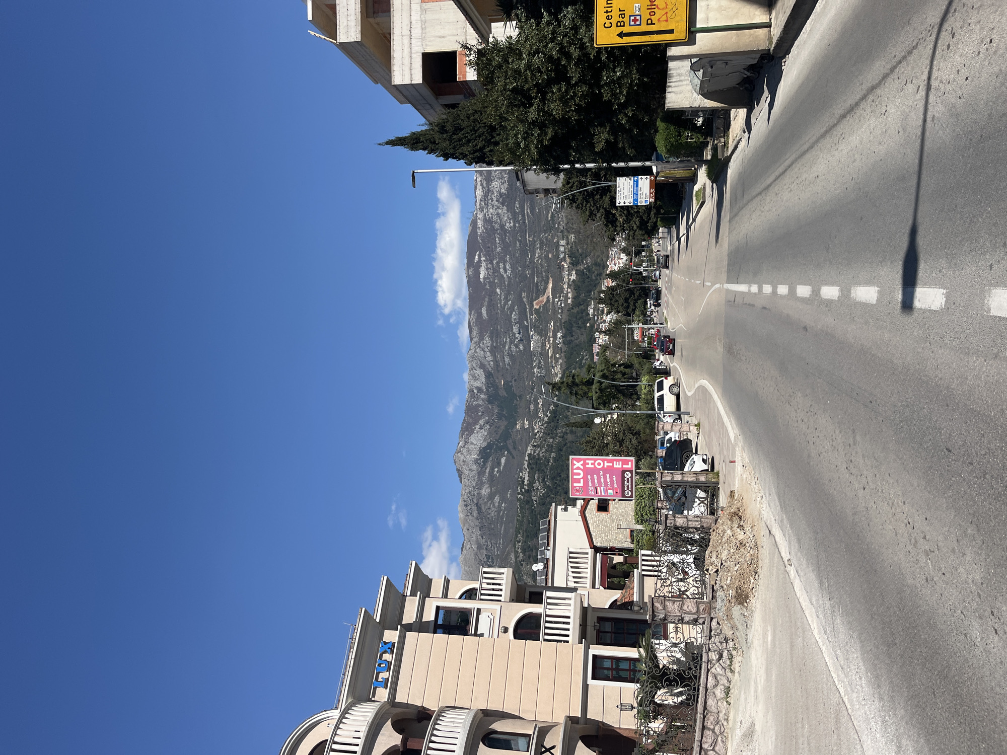 The image depicts a street in Budva, Montenegro, with a view of mountains in the background. The sky is clear blue. On the right side of the road, there are several cars parked, as well as a sign for the "Lux Hotel" visible on the right. A street lamp is seen with a shadow cast across the road. Traffic and pedestrian crossing are also visible in the distance. There are buildings on both sides of the road, with some under construction. The road has white dashed lines marking the center. There is also a signpost with destinations to Cetinje, Bar and Police. The buildings are beige.