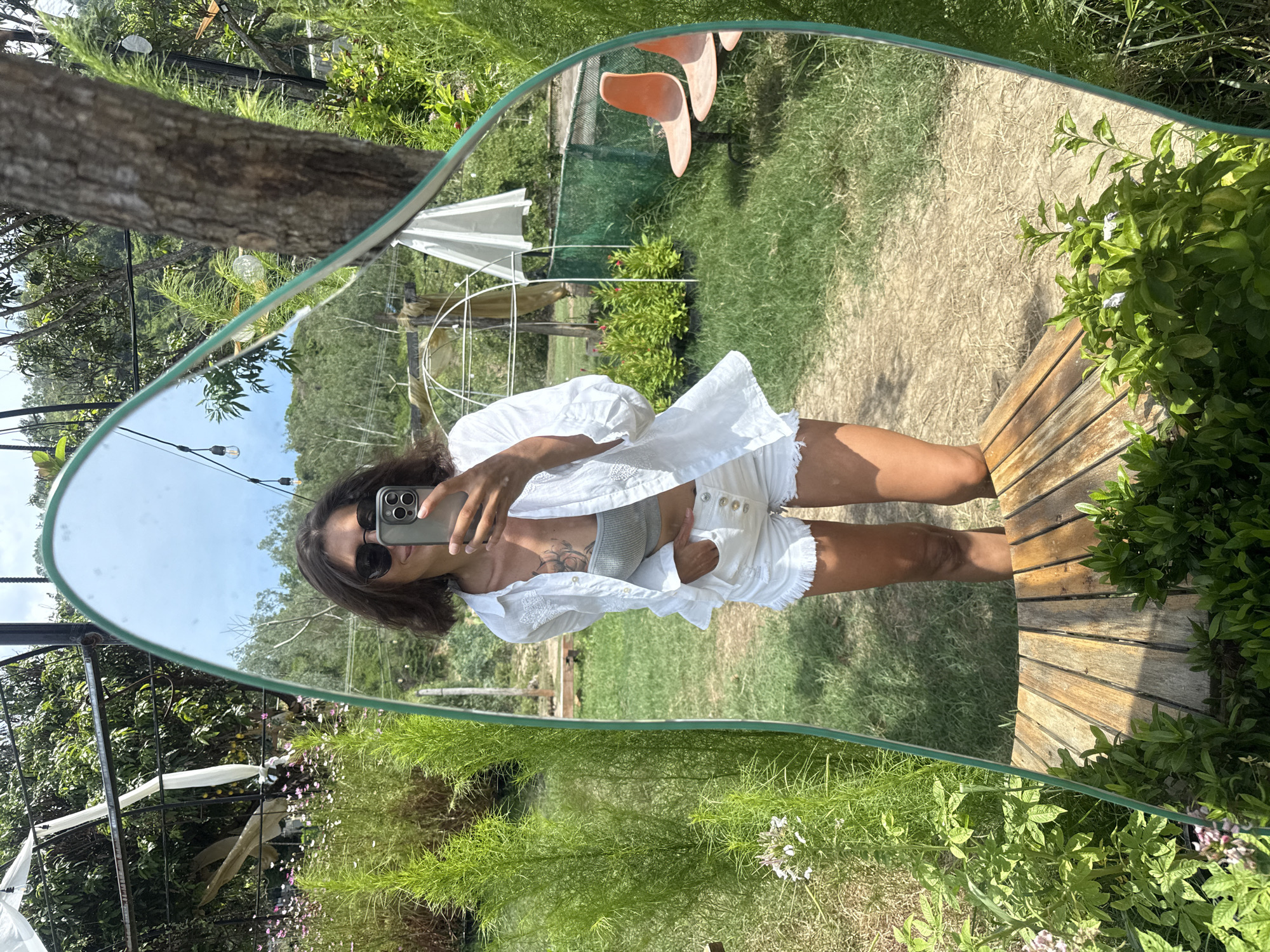 A woman is taking a selfie in an ornate mirror placed outdoors amidst lush greenery. She is wearing sunglasses, a white open-collared shirt over a grey crop top, and white denim shorts with frayed edges. Her left hand is tucked into her pocket. The mirror reflects the woman, the surrounding garden, and the sky. In the background, there are trees, flowers, a fence, and two orange chairs. The scene suggests a relaxed, outdoor setting, possibly a cafe or garden in Vĩnh Lương, Vietnam.