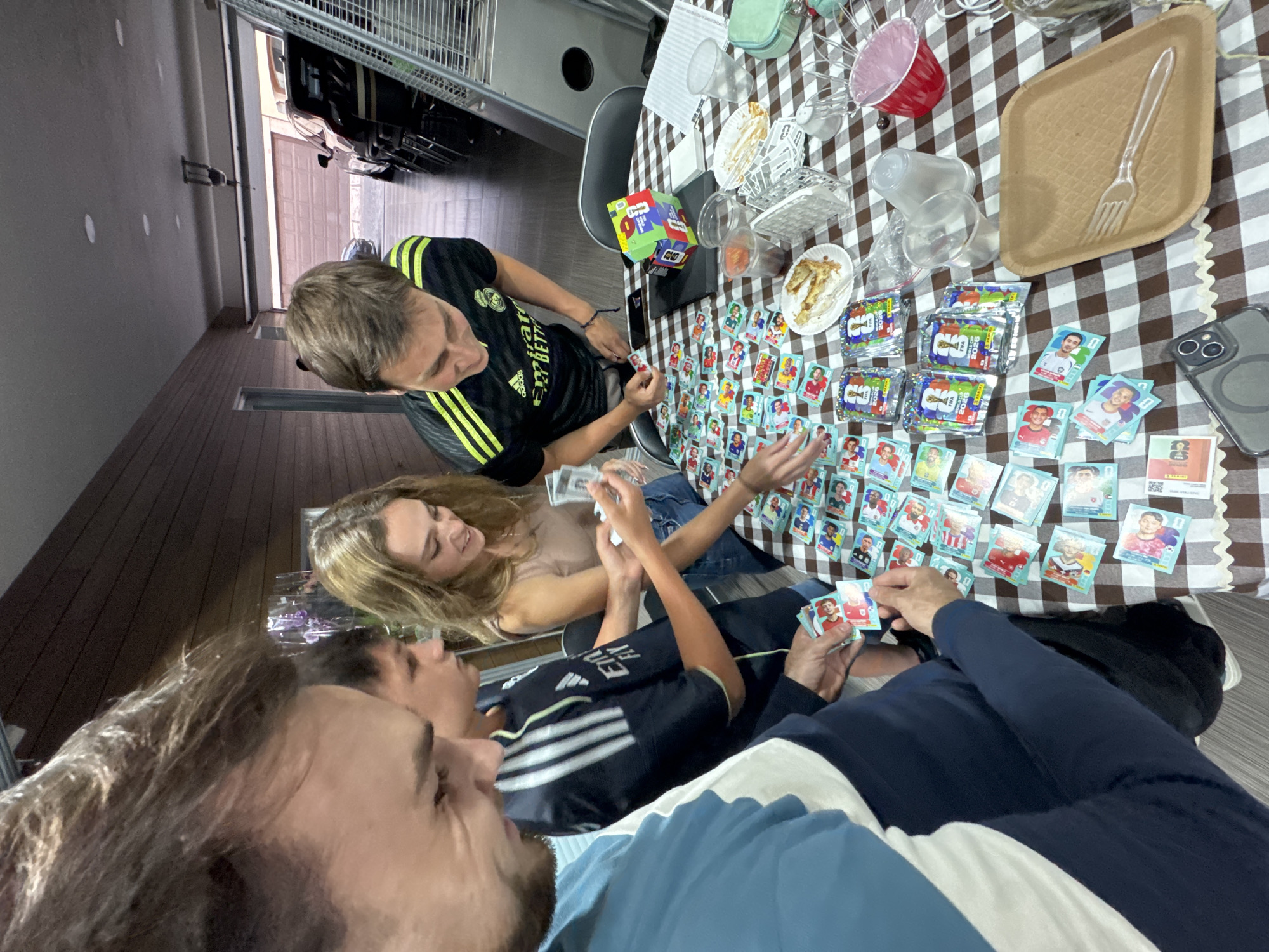 A top-down shot captures a group of four people gathered around a table covered with a brown and white checkered tablecloth. They are sorting and examining FIFA World Cup 2026 sticker packs. Scattered around the table are opened packs of stickers, loose stickers, disposable plates, cups, and other containers. One person is wearing a black Real Madrid jersey with yellow stripes, and another is wearing a black jersey with white stripes. A cell phone rests on the table. The location is San Bernardino Tlaxcalancingo, Mexico.