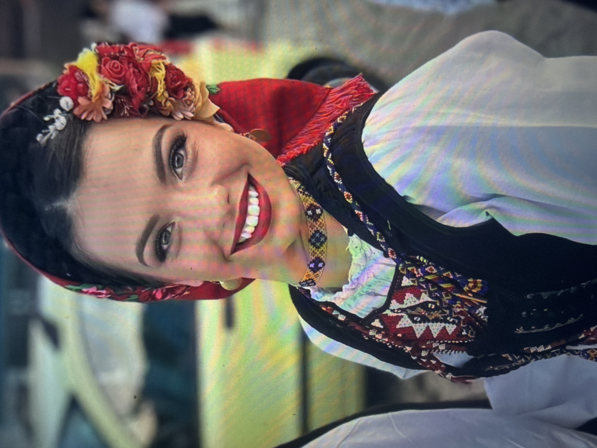A young adult woman is depicted in a close-up, chest-up portrait, smiling broadly in an outdoor setting in Shumen, Bulgaria. She has dark hair, dark eyes, and is wearing red lipstick, revealing white teeth in a wide smile. Her head is adorned with a dark red headscarf featuring subtle floral patterns, and a prominent cluster of red, yellow, and orange artificial flowers is secured on the right side of her head. She wears gold-toned, circular earrings. Around her neck is a choker-style necklace with a dark blue and gold/yellow beaded geometric pattern. Her attire consists of a white, long-sleeved shirt with ruffles at the neckline, layered under a dark-colored vest. The vest is intricately embroidered with traditional geometric patterns in red, white, dark blue, yellow, and orange, and features braided decorative elements. The background is significantly blurred, showing a light yellow vehicle to the left and indistinct shapes suggesting other people or structures under daylight conditions.