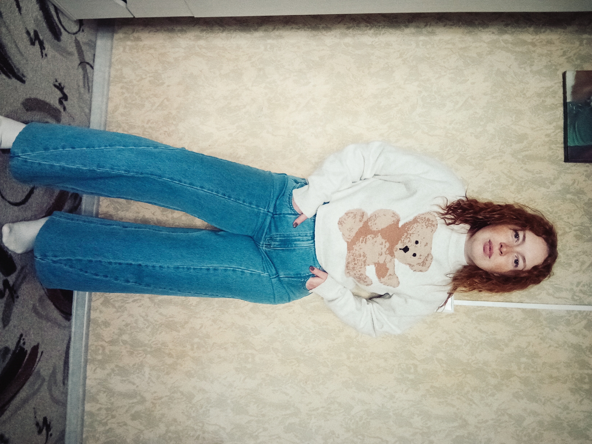 A young person with reddish-brown, curly hair is seen lying on their back, angled diagonally across the frame, looking directly at the camera. They are wearing a white long-sleeved turtleneck sweater adorned with a large brown teddy bear graphic on the front. Their attire also includes light blue, wide-leg denim jeans and white socks. Their hands are positioned on their hips.
The setting is indoors, featuring two distinct floor or wall surfaces. To the left, there is a grey carpet or rug with abstract black and dark brown patterns. The larger area beneath and around the person is covered by a cream or beige-colored surface with a subtle, swirling pattern. A thin, light-colored strip separates these two patterned areas. A dark, possibly framed, object is partially visible in the upper right corner. The person displays a neutral expression. No visible text is present in the image. This scene is located in Yurgamysh, Russia.
