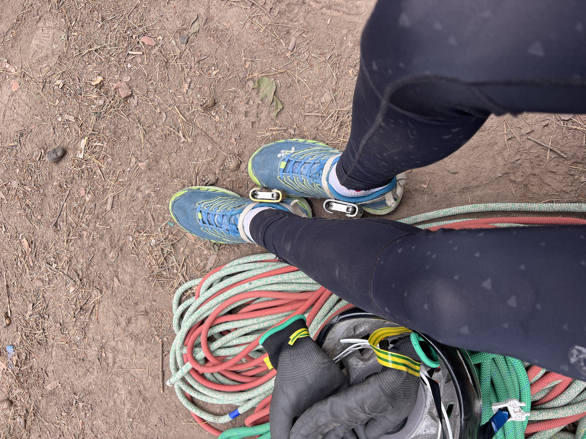 The image shows a person's legs and feet from a top-down perspective, resting on the ground. The ground is covered with dirt, small twigs, and dry leaves, consistent with an outdoor environment. The person is wearing black leggings with a subtle geometric pattern and blue athletic shoes with lime green accents. Attached to the laces of each shoe is a carabiner, one appears to be brass-colored and the other silver.
To the left of the legs is a pile of climbing ropes, a helmet, and a pair of gloves. The ropes are coiled and appear to be red and green. The helmet is black with yellow and green straps. The gloves are dark grey.
The location is identified as Chaikovsky, Russia, a city situated on the Kama River. This environment, with its natural ground cover and climbing equipment, suggests an outdoor recreational area, possibly near a climbing or outdoor adventure sports facility within or on the outskirts of Chaikovsky.