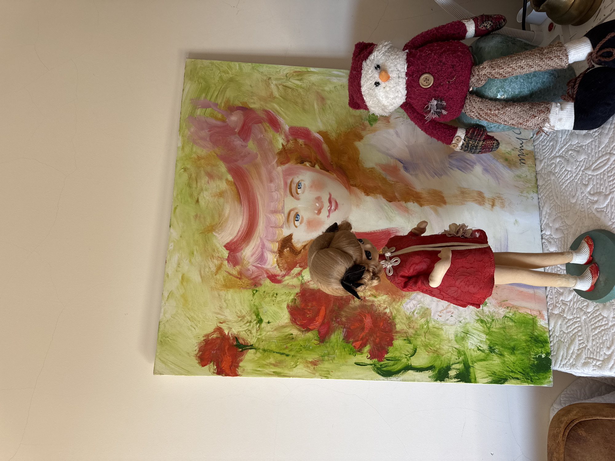This image depicts an indoor scene in Mirandela, Portugal, featuring a collection of decorative items arranged on a white, textured tablecloth. Dominating the background is a large, rectangular, impressionistic oil painting leaning against a pale beige wall exhibiting minor hairline cracks. The painting portrays the bust of a woman with light reddish-blonde hair and blue eyes, wearing a broad-brimmed pink hat, set against a vibrant green background adorned with several prominent red floral elements. A dark, cursive signature is visible in the lower right corner of the canvas.
In the foreground, two figures are positioned in front of the painting. To the left, a doll with blonde hair styled in two buns, adorned with small black bows, stands on a dark green circular base. The doll wears a textured red dress with a white decorative knot closure at the neckline, white socks, and red shoes. To the right, a soft, fabric snowman figure is seated atop a teal ceramic vase or pot. The snowman has a white textured head, an orange carrot-shaped nose, black bead eyes, and a stitched mouth. It is dressed in a dark red hat and body, featuring a large brown button on its chest and smaller buttons on its mittens, which are patterned in brown and plaid. Its legs are made of brown speckled fabric, terminating in white socks and black shoes. Partially visible in the lower left corner is the brown upholstered back of a chair, and in the lower right, a brass-colored pot or bowl.