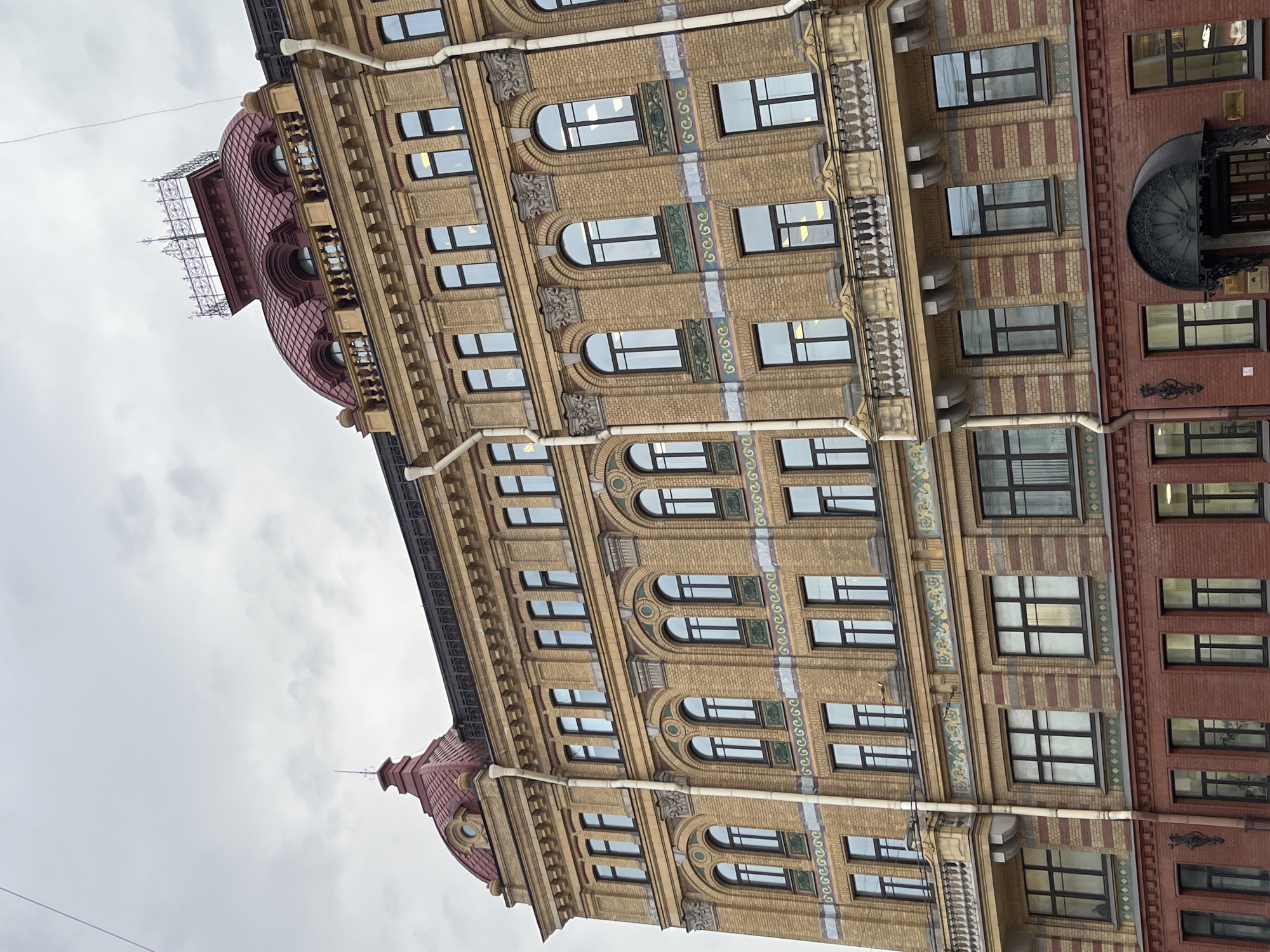 The image shows the exterior of a building in Unknown, Unknown. The building features multiple stories with numerous windows. The architecture is detailed, with decorative elements along the façade. There's a distinct color variation between sections of the building's brickwork. Above, the sky is overcast.