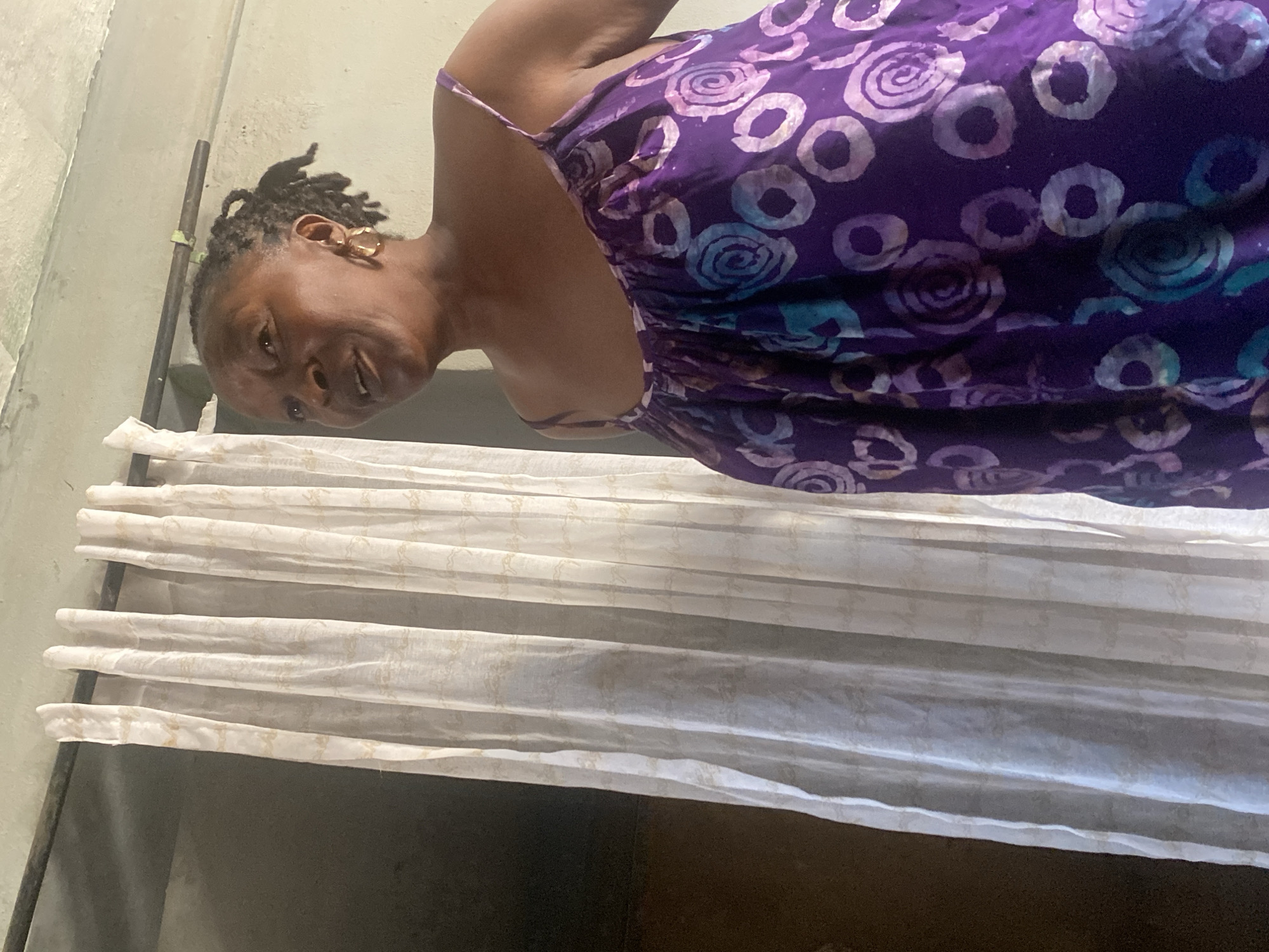 A woman with dark braided hair and gold hoop earrings is the central subject of this indoor portrait, captured in Sabon Tasha, Nigeria. She looks directly at the camera with a faint, pleasant smile. She is wearing a vibrant purple spaghetti-strap dress featuring an intricate pattern of white and light-blue circles and spirals, resembling a batik or tie-dye design.
The setting appears to be a casual interior space, possibly a home. Behind the woman is a light-colored wall, which could be concrete or painted. To her left, a dark, slender vertical rod or pipe is partially visible against the wall. The lower portion of the image shows pleated white or off-white curtains or blinds, displaying a subtle, light-colored pattern. Below the curtains, there is a dark, undefined area, possibly a floor or dark furniture. The natural light suggests the photograph was taken during the daytime. No visible text is present in the image.