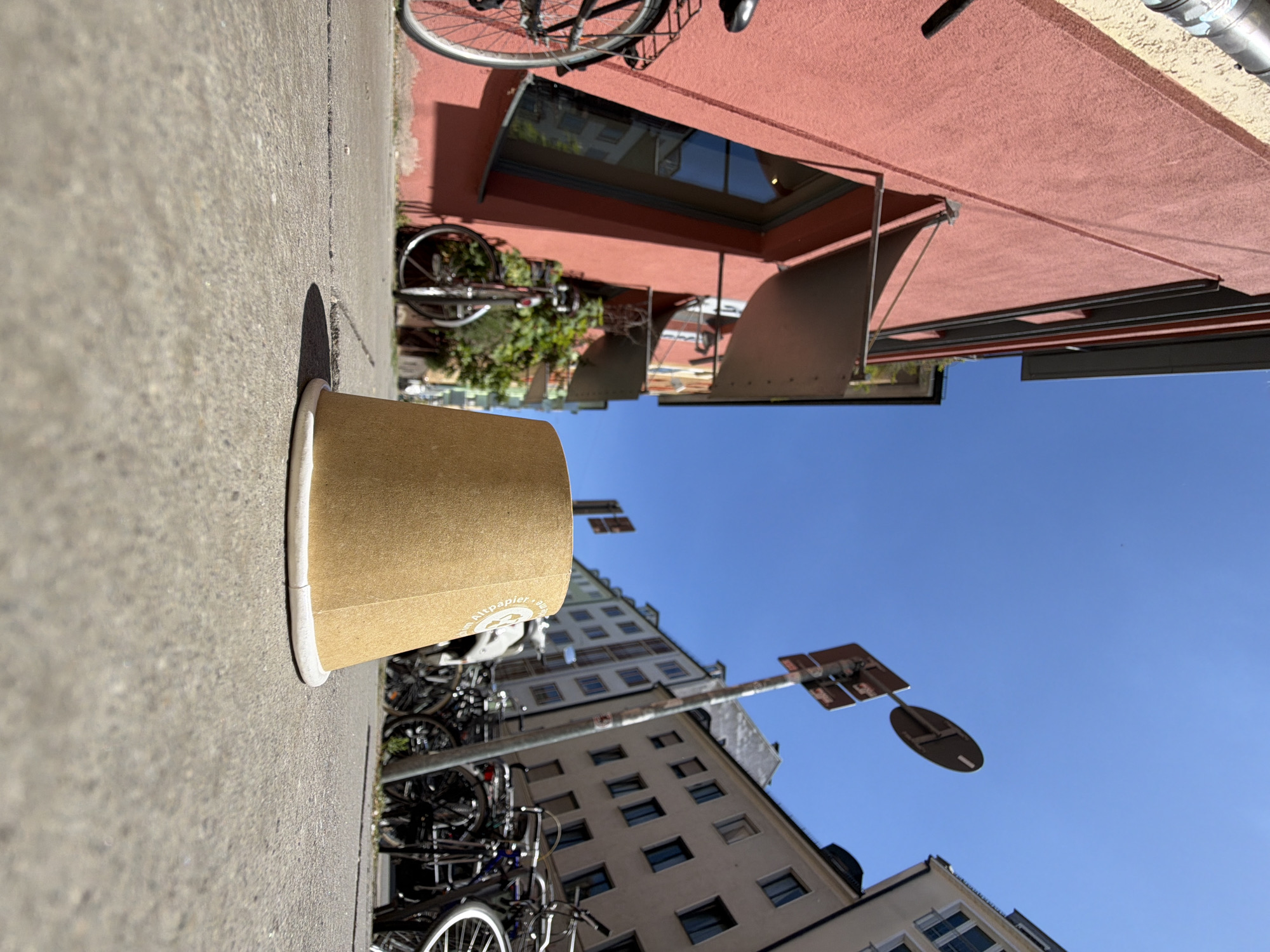 The image captures a street scene in Munich, Germany, taken from a low, ground-level perspective. In the foreground, a single disposable paper cup, light brown with white rim and some faint text, rests on a concrete sidewalk. The cup is positioned slightly off-center to the right. Behind the cup, the sidewalk stretches into the background, showing some cracks and texture.
To the left of the cup, a bright pink building dominates the scene, its textured facade illuminated by sunlight. A metal awning extends from the building over a dark-framed window. A black bicycle with a rear rack is partially visible on the far left, its front wheel cut off by the frame. Further back along the building's facade, another bicycle is parked, its front wheel and handlebars visible.
In the background, a row of multi-story buildings lines the street, their facades painted in various light colors and featuring numerous windows. A metal street signpost stands near the center of the background, with two signs attached, one a circular road sign and the other a rectangular sign with some text. A cluster of bicycles is parked along the right side of the street, their wheels and frames forming a dense arrangement. The sky is a clear, bright blue, indicating a sunny day. The overall impression is of a typical urban street with everyday objects and infrastructure.