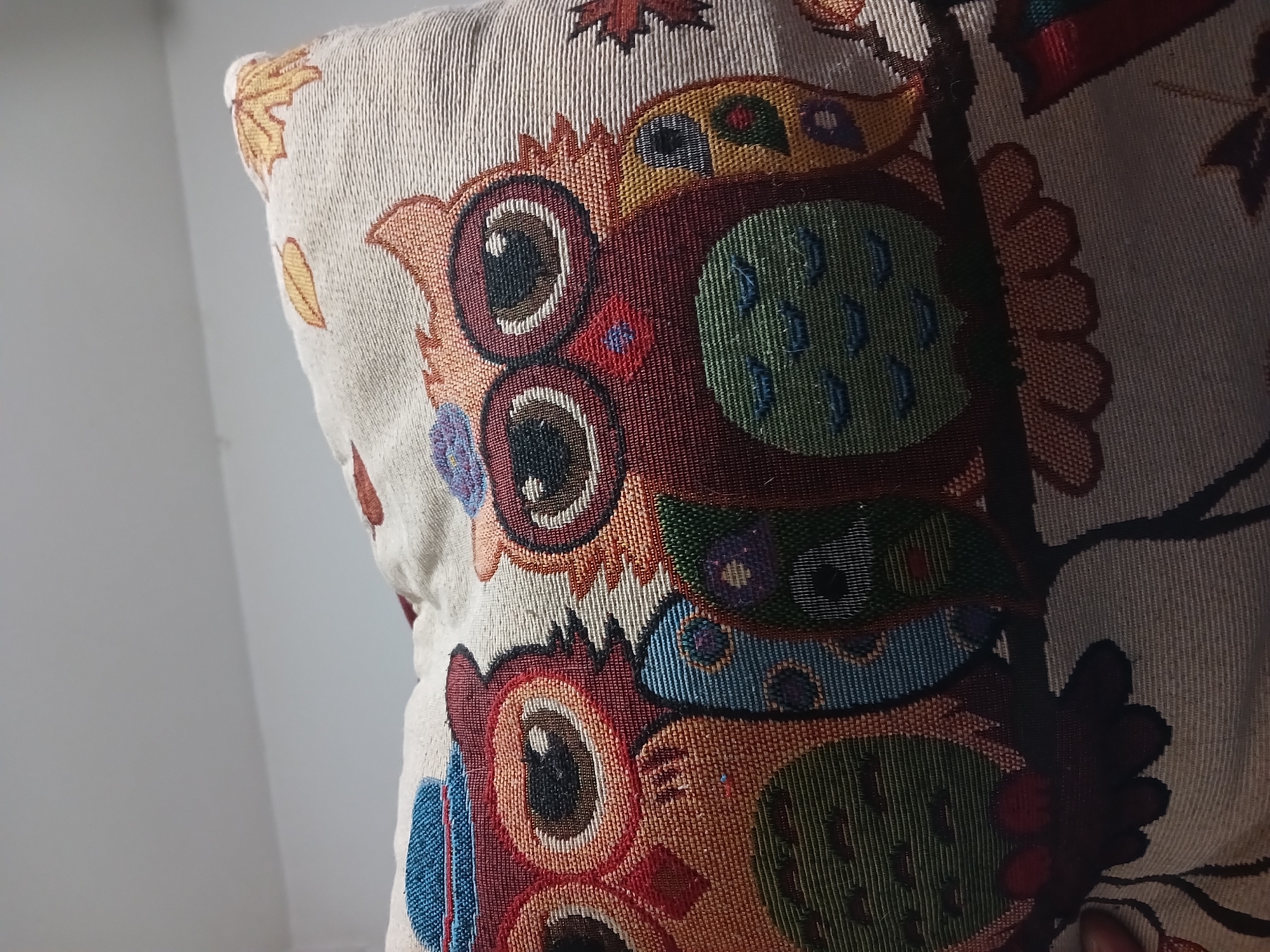 A close-up shot of a pillow with a woven owl design. The pillow has a beige background with colorful owls and maple leaves. The pillow is in front of a white wall and illuminated by sunlight from the left. There is no indication of the city of Buenos Aires, Argentina.
