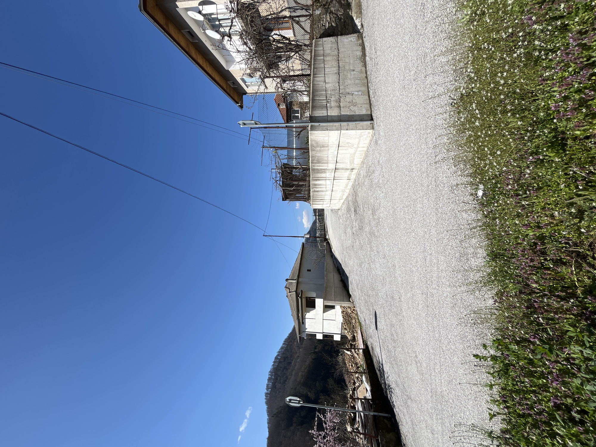 The image depicts a street scene in Srednogortsi, Bulgaria, observed on a clear, sunny day. The angle is looking down a narrow asphalt road that is bounded on both sides by adjacent buildings and vegetation.
On the right side of the road, there is lush vegetation with green foliage and small white and purple flowers, partially obscuring the lower edge of the buildings.
The buildings are constructed from concrete blocks. A series of satellite dishes are affixed to one of the buildings.
The road itself is grey.
Powerlines run above the buildings and across the blue sky. The sky is clear with no clouds. In the background, a tree-covered hillside is visible.
A road sign can be seen on the lower part of the left side of the image.
