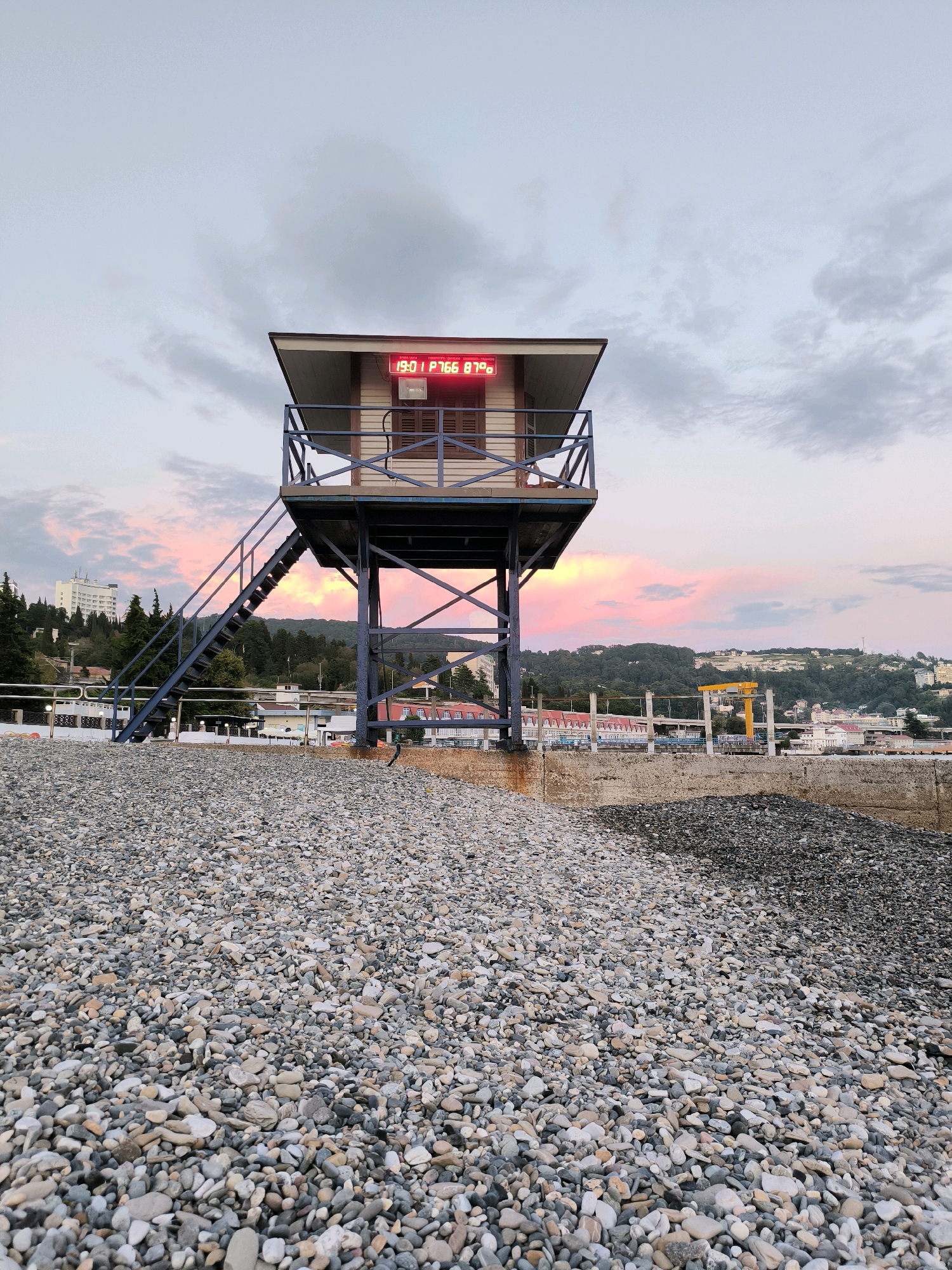 An eye-level, wide shot captures a rocky beach in Sochi, Russia, during twilight. A lifeguard tower, painted white with a brown roof and blue railings, stands prominently in the foreground, elevated on dark metal stilts. A blue metal staircase ascends to the tower's platform. Above the entrance to the tower, a red digital display shows "19:01 P766 87°C". Behind the tower, a cityscape unfolds with trees and buildings nestled on a hillside, bathed in the soft hues of a pink and orange sunset sky. The beach itself is covered in smooth, grey and multicolored pebbles.