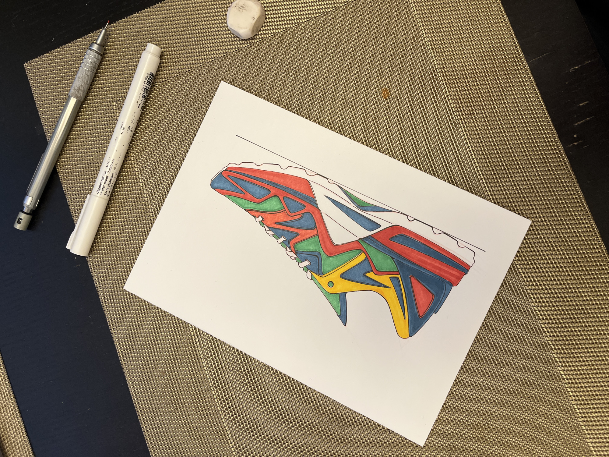 A drawing of a shoe is placed on a white piece of paper. The drawing is outlined in black ink and colored with blue, red, green, and yellow markers. Above the drawing, on a woven beige mat, are a mechanical pencil, a white marker, and a white eraser. Part of a black table is visible on the left side of the image.