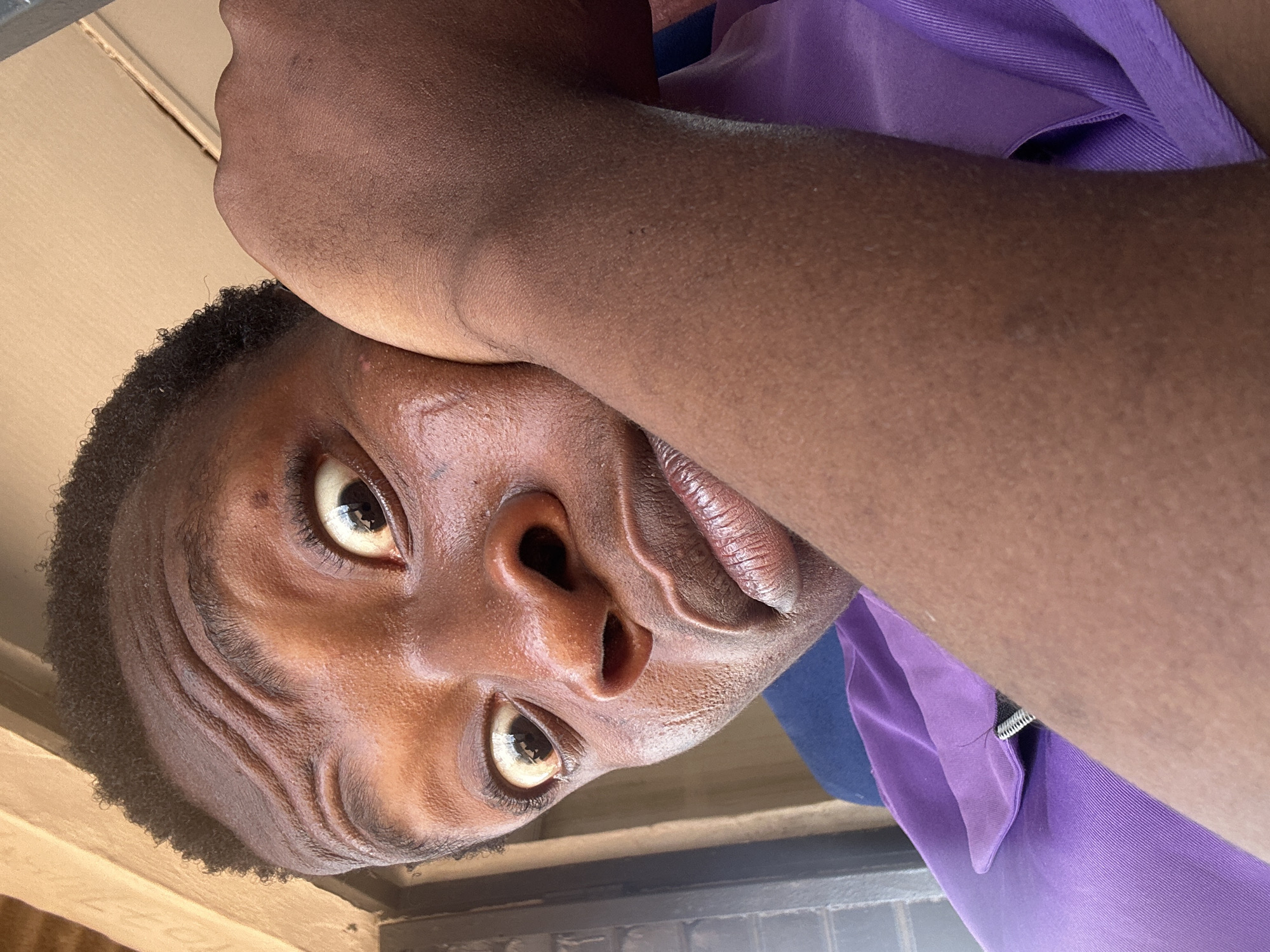A close-up, tilted selfie of a young Black male with short, dark hair. His left forearm rests on his forehead. He wears a purple collared shirt. The location appears to be indoors.