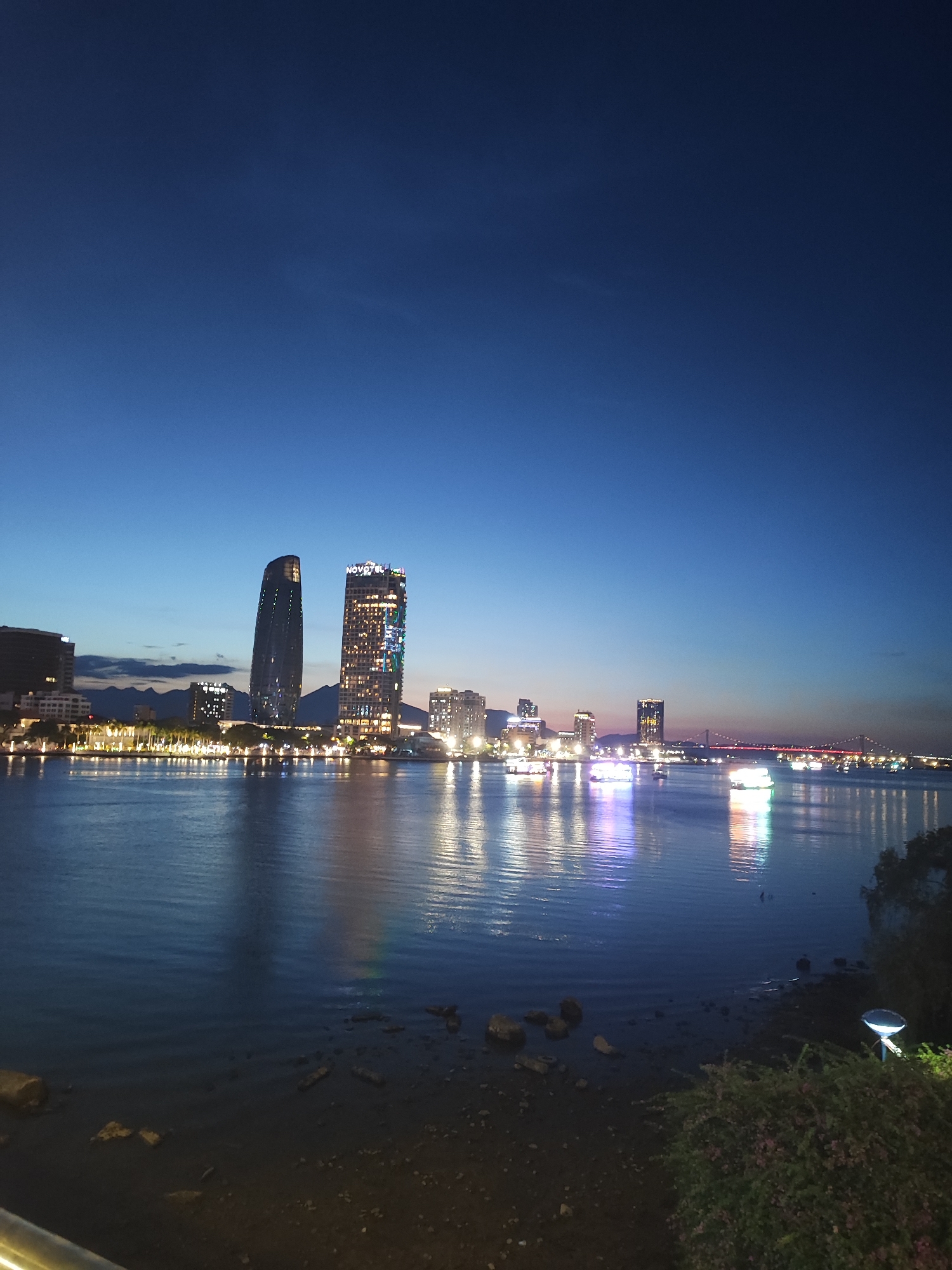 This is a nighttime shot of the city of Đà Nẵng, Vietnam, from across the Hàn River. The water dominates the bottom portion of the image, reflecting the lights of the city. Several buildings and structures appear illuminated along the riverfront, most prominently the Novotel Danang Premier Han River hotel and the taller buildings to its left. In the background, faint hills can be seen. The sky is a gradient of dark blues. The foreground shows a small section of the riverbank with dark earth, rocks, and some vegetation to the right. A lamp is on the right.