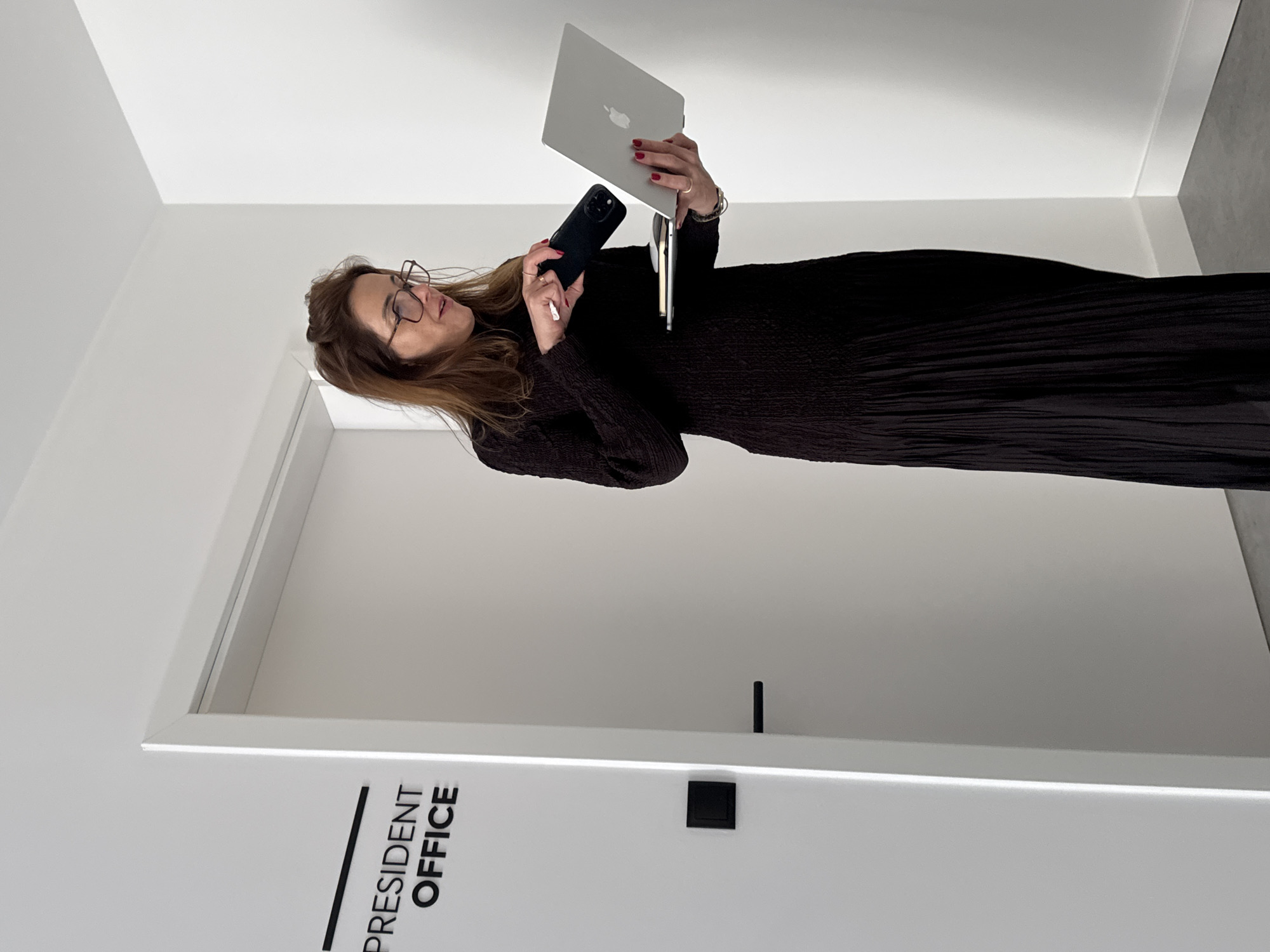 The scene is a modern, minimalist office hallway in Torres Vedras, Portugal, characterized by white walls and a gray floor. A sign on the left white wall reads "PRESIDENT OFFICE" in black capital letters with a black underline. A black square panel is mounted below the sign, and a white door with a black horizontal handle is partially visible.
A female subject stands centrally, facing slightly to her right. She has brown hair, wears eyeglasses, and is dressed in a dark, textured, long-sleeved floor-length dress. Her fingernails are painted red, and she wears rings on her left hand and a bracelet on her left wrist.
In her left hand, she holds an open silver MacBook laptop, an Apple logo visible on its lid. Balanced on the laptop or a surface beneath it are a white wireless mouse and a small notebook. Her right hand holds a dark-cased smartphone, which she appears to be operating or cleaning with a small white tissue held between her thumb and forefinger. Her gaze is directed downward towards the devices, and her mouth is slightly open, suggesting focus or speech.