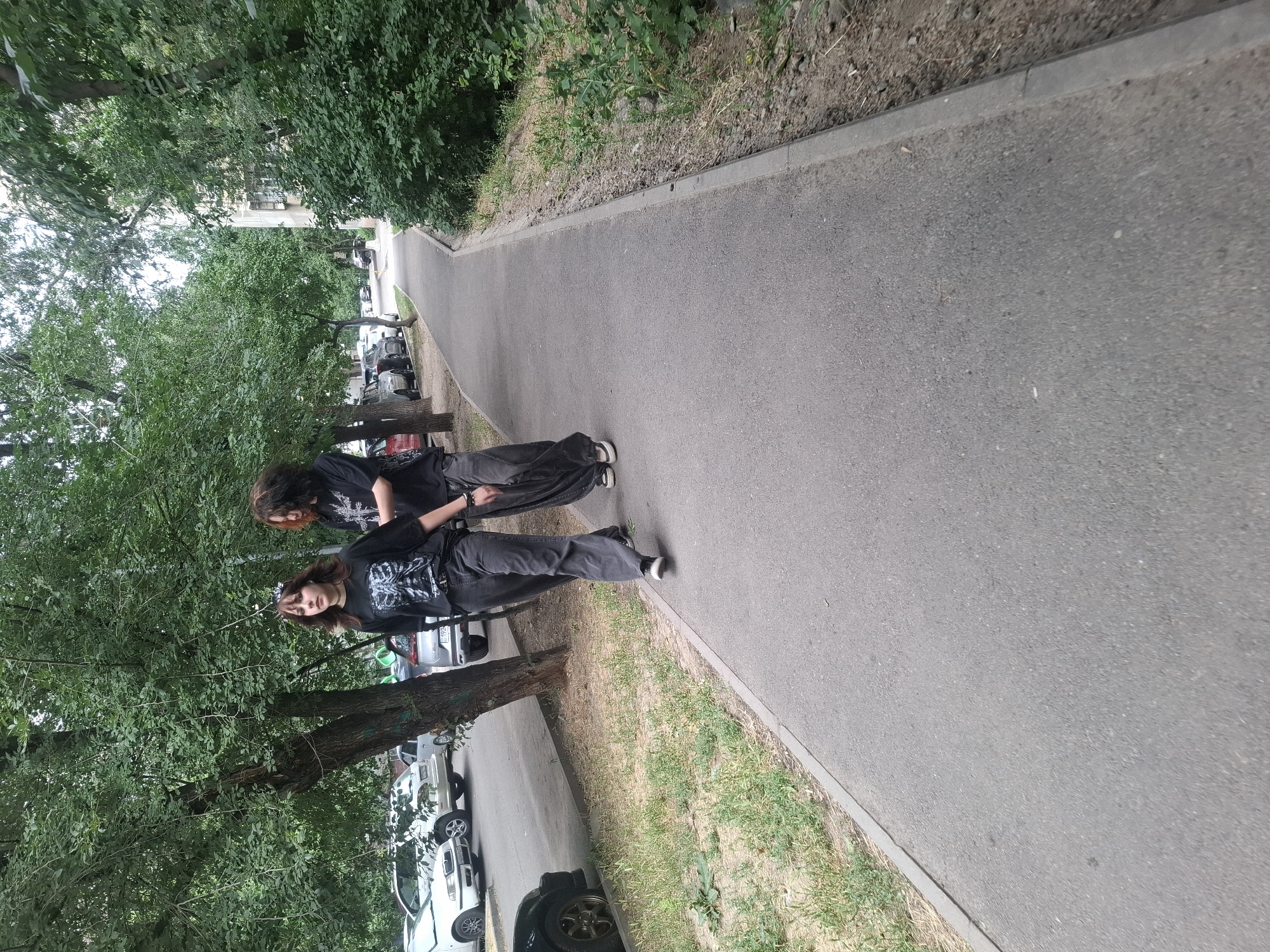 Two individuals, one with long dark hair and a black t-shirt with a graphic print, and another with darker, shorter hair and a black t-shirt with a skeletal print, walk along a paved pedestrian path. They are both wearing dark, loose-fitting pants and dark sneakers. The path runs alongside a grassy area with some weeds and is bordered by a curb. Beyond the grassy area, there is a row of parked cars, including a white Skoda. A large tree with dense green foliage is to the left of the path. Further in the background, there are more trees and a multi-story building. The scene appears to be an urban residential area. The provided hints suggest this scene is located in Almaty, Kazakhstan.