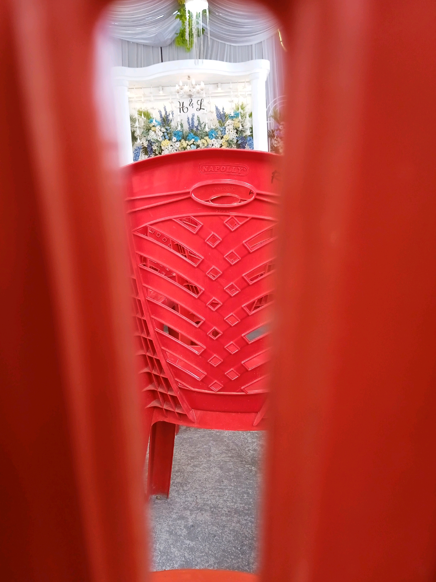 A red plastic chair is positioned in the foreground, partially obscuring a wedding decoration. The wedding decoration features white draping, flowers in blue and white hues, and a chandelier. The letters "H & L" are prominently displayed on the decoration. The chair has the brand name "NAPOLLY" embossed on its back. The setting appears to be an indoor venue in Kelurahan, Indonesia, possibly decorated for a wedding ceremony.