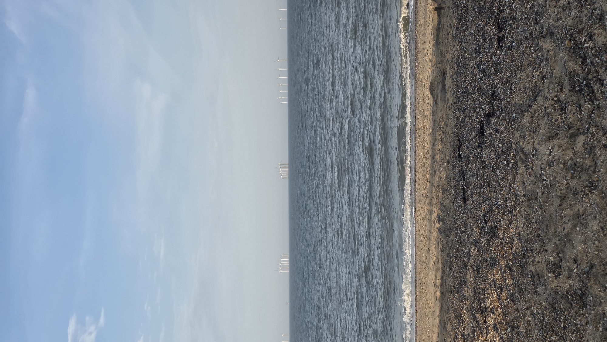 The image depicts a wide, panoramic view of a windswept coastline. The scene is divided into three distinct horizontal bands. The leftmost band is dominated by a pale blue sky with wispy white clouds, suggesting fair weather. The middle band is the vast expanse of the sea, its surface rippled with gentle waves. In the distance, a line of wind turbines stands tall on the horizon, hinting at an offshore wind farm. The rightmost band shows the beach, a mix of sand and pebbles, with a gentle surf washing ashore. The overall impression is one of natural beauty and industrial presence, with the vastness of the sea and sky contrasting with the man-made structures. The lighting suggests it is daytime, likely mid-morning or late afternoon, given the soft light. There is no visible text in the image.