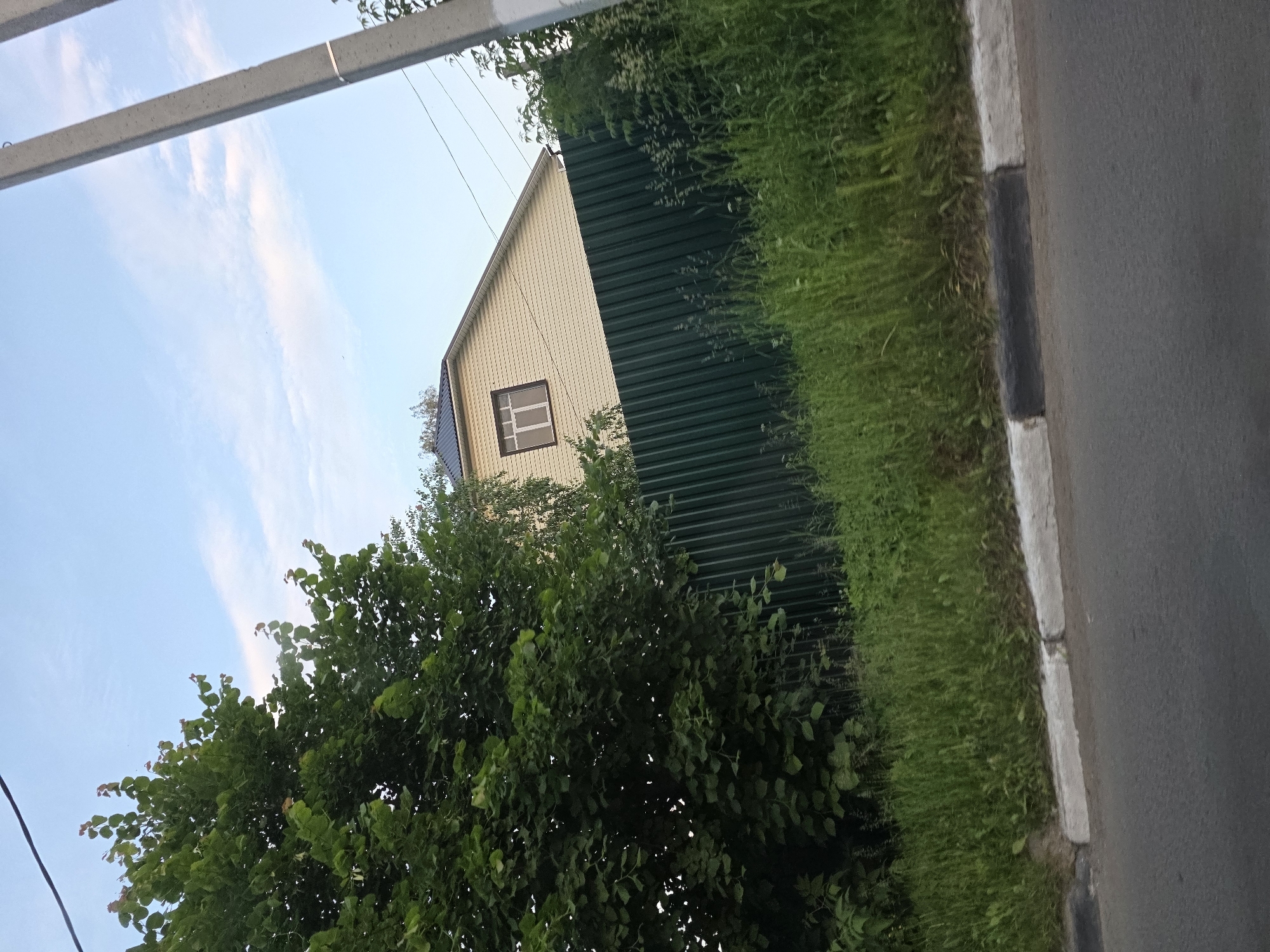 A wide shot captures a scene in an outdoor urban environment with a clear sky and scattered clouds. The foreground features a paved road with a concrete curb and an adjacent patch of tall green grass. Beyond the grass is a dark green corrugated metal fence that stretches across the frame. Partially visible behind the fence, to the right, is a yellow house with a dark roof and a single small window. To the left of the house, a large green tree with dense foliage dominates the left side of the image, partially obscuring the fence and the house. A gray utility pole stands on the right side of the image, positioned behind the fence and next to some greenery. The lighting suggests it is daytime, possibly late afternoon or early evening.