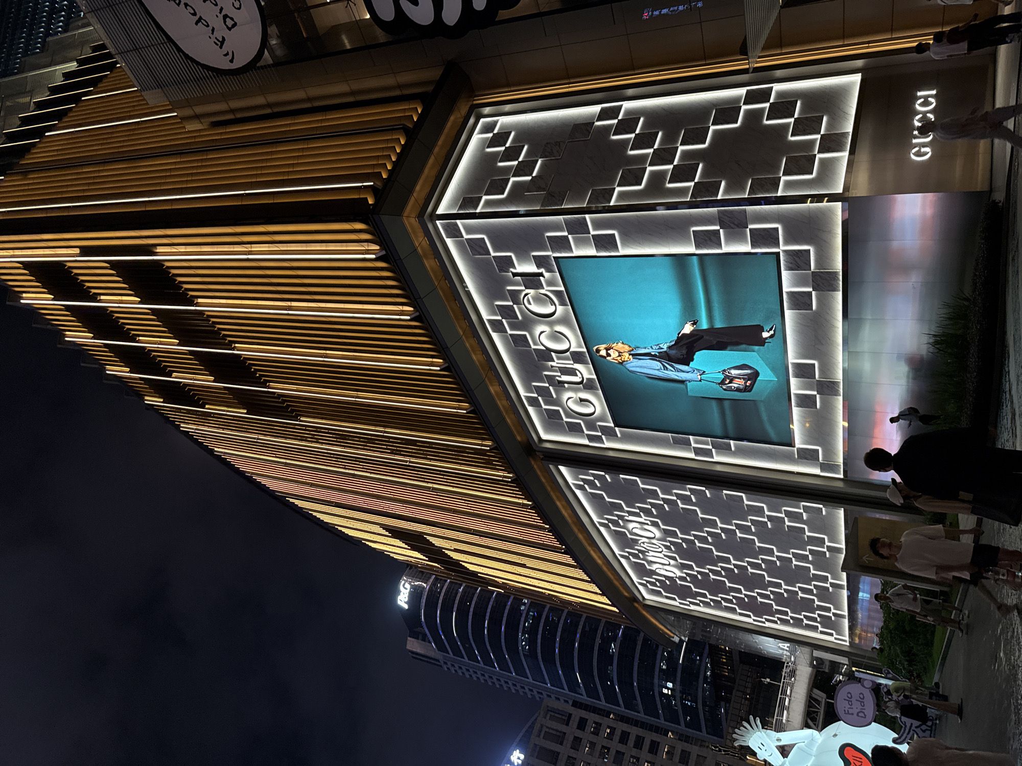 The image depicts a nighttime scene outside a modern Gucci store in Tianhe District, China. The building features an illuminated facade with geometric patterns and a large digital screen displaying a fashion advertisement with a person reclining with a suitcase. The store's name, "GUCCI," is prominently displayed in glowing letters.
Several people are visible, some standing and observing the storefront, while others walk past. The lighting is a mix of warm yellow from the building's exterior design and the cool glow from the digital screen and interior lights. A large, inflatable cartoon figure is also present in the foreground, adding a whimsical element to the urban environment. The overall impression is one of a lively commercial district at night, with a focus on luxury retail and consumer activity. Some visible text includes "GUCCI" on the building and "Fido Dido" on the inflatable figure.