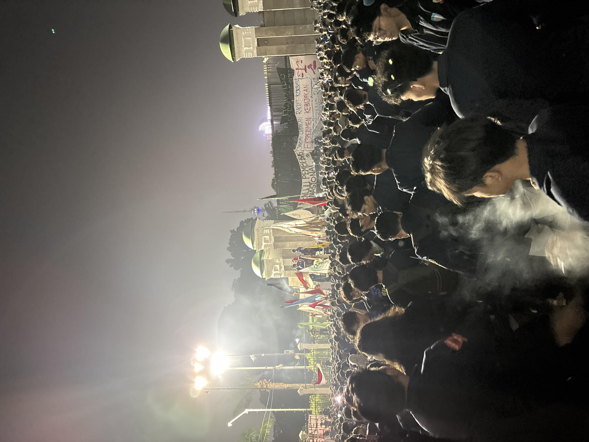 The image depicts a nighttime gathering of a large crowd of people. The orientation of the photo is portrait, with the scene extending upwards. In the foreground, the heads and shoulders of individuals are visible, many wearing dark clothing. Smoke or fog is present, creating a hazy effect around the people.
Further into the scene, a banner is unfurled, held by some of the individuals. The banner has text, but it is largely obscured by the crowd and the angle. Some legible phrases on the banner include "JANGAN JADIKAN RAKYAT KORBAN," "EKSPERIMEN KEBIJAKAN NASIONAL," and "TINGKATKAN KUALITAS HIDUP BANGSA." In the background, a building with domes and pillars is visible, illuminated by artificial lights. Several flags are also present, adding color to the scene. The overall atmosphere appears to be one of a rally or protest, given the banner and the large assembly of people. The lighting suggests it is nighttime, and there are strong spotlights contributing to the illumination. The location cues indicate Central Jakarta, Indonesia.