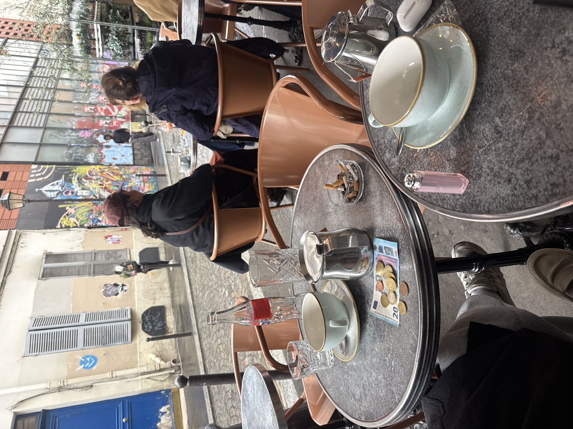 The image captures an outdoor scene in Paris, France, likely in a cafe or bistro setting. Two tables are visible, made of gray stone with metal rims. Each table has a tea cup and saucer, along with various objects. One table has a lighter and an airpods case; the other has a tip of Euro coins and a 20 Euro bill. An ashtray with cigarette butts sits on the table next to the tips. A sugar dispenser, glass bottle and water glass are also on the table. A shiny metal tea pot is on the table with the money. Several orange chairs are situated around the tables, with people seated in them. The attire of the people suggests casual wear. In the background, the architecture indicates a European urban environment, with buildings and graffiti visible. The image is taken from a high-angle perspective, with a slightly tilted view.