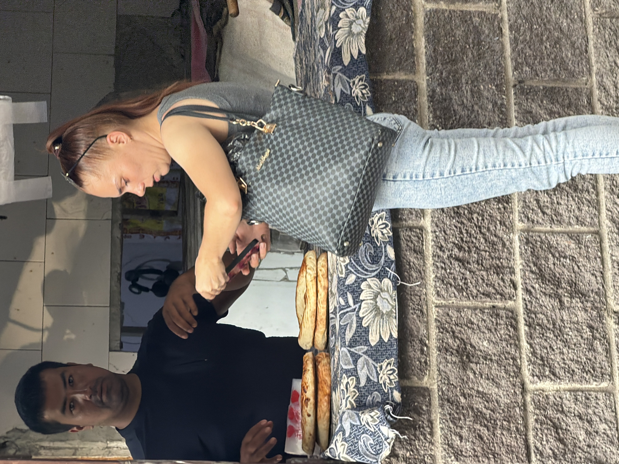 A man in a black t-shirt stands behind a table laden with bread, facing a woman in a grey tank top and light blue jeans. The woman is looking down at her phone, which she holds in her right hand, and has a black checkered handbag slung over her left shoulder. The table is covered with a blue and white floral patterned cloth. The background consists of a brick wall and what appears to be a market stall. The scene is outdoors.