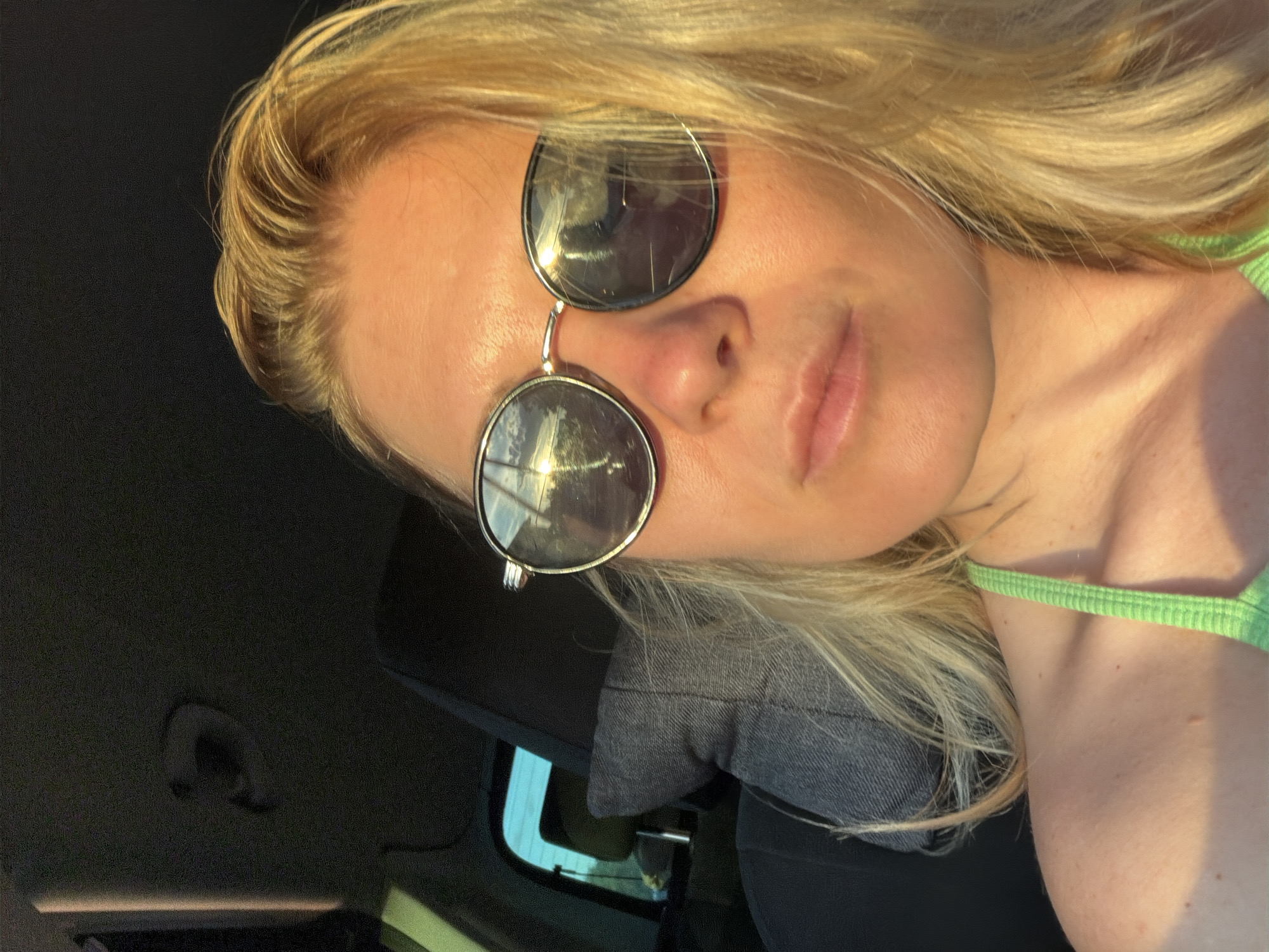 A woman with blonde hair and sunglasses is sitting in the passenger seat of a car. The sun is setting, casting a warm glow on her face. The car's interior is visible in the background, with a headrest and the car's ceiling. The woman is wearing a green top. The image appears to be a selfie taken in the city of Aleksandrovskoye, Russia, during golden hour.