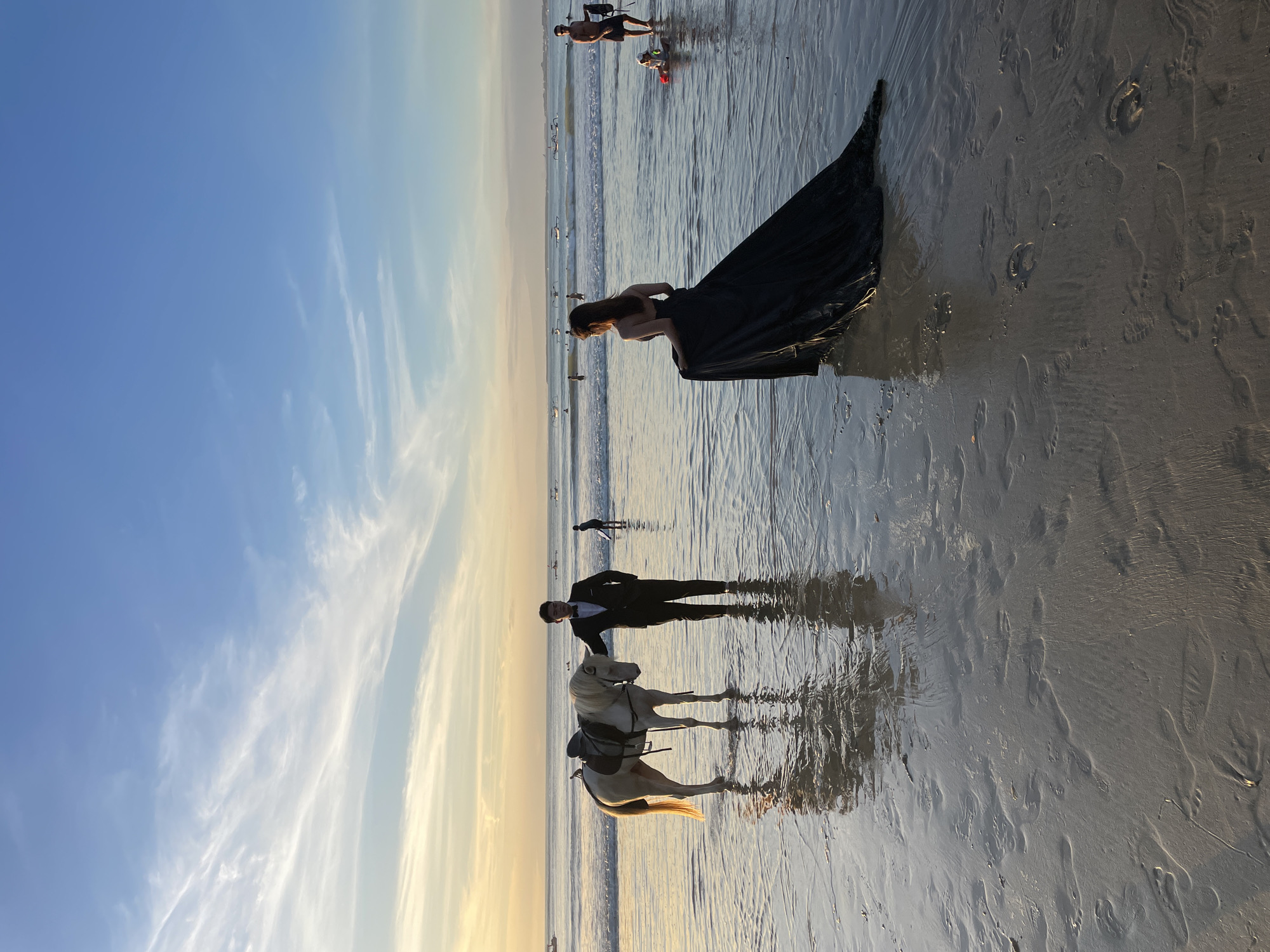 The image shows a beach scene in Unknown, Unknown. In the foreground, a man in a black tuxedo stands next to a white horse with a saddle. Behind them, further into the water, a woman in a long, dark dress stands facing away from the camera. In the background, a few other people are visible in the water near the shoreline. The sky is a clear blue with wispy clouds, and the sun appears to be setting, casting a warm glow on the sand and water. Reflections of the subjects and the sky are visible on the wet sand.