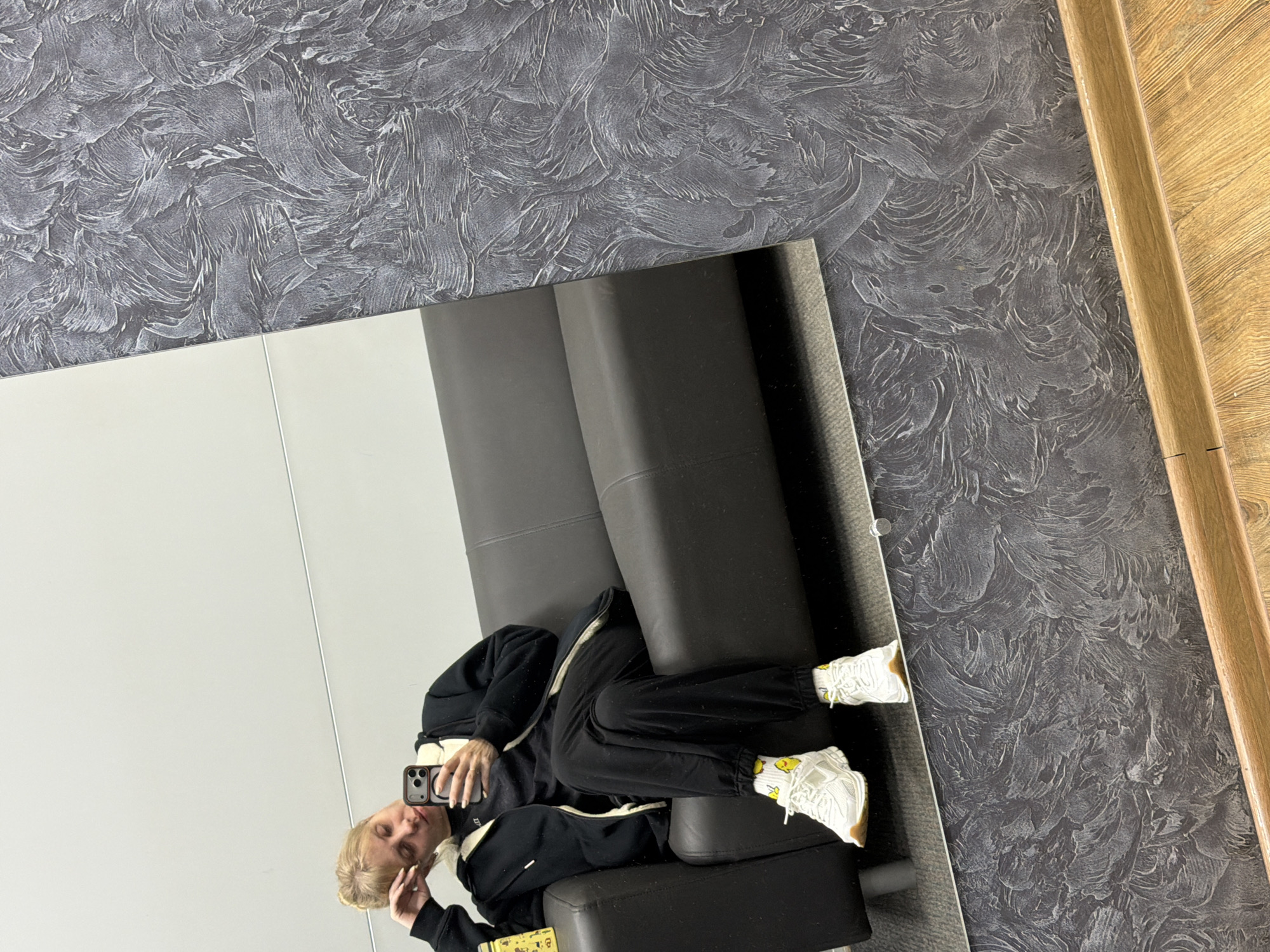 A reflection in a mirror shows a person lying on a dark gray couch in a room. The person has blonde hair and is wearing a black and white tracksuit with white sneakers and yellow socks. They are holding a phone and taking a selfie. The room has a textured gray wall and a wooden trim. The location is not explicitly stated as Chaikovsky, Russia, but is inferred based on image metadata.