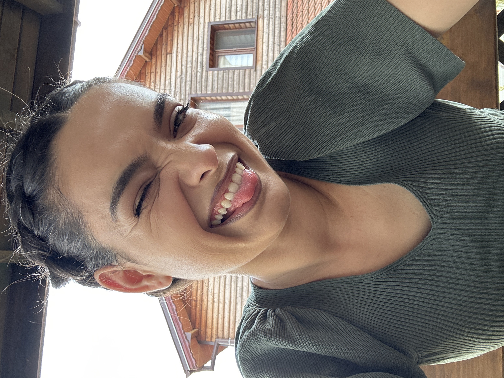 A young woman with braided dark hair is shown in a close-up selfie. She is winking and sticking her tongue out at the camera, with a wide, playful smile. Her skin appears smooth and lightly tanned. She is wearing a dark olive green ribbed knit top with puffed sleeves. The background consists of a wooden building with a sloping roof and a window, suggesting an outdoor setting. The overall impression is one of a casual and cheerful moment captured on camera. The location is specified as Malevo, Bulgaria, though no specific city landmarks are visible in the image itself.