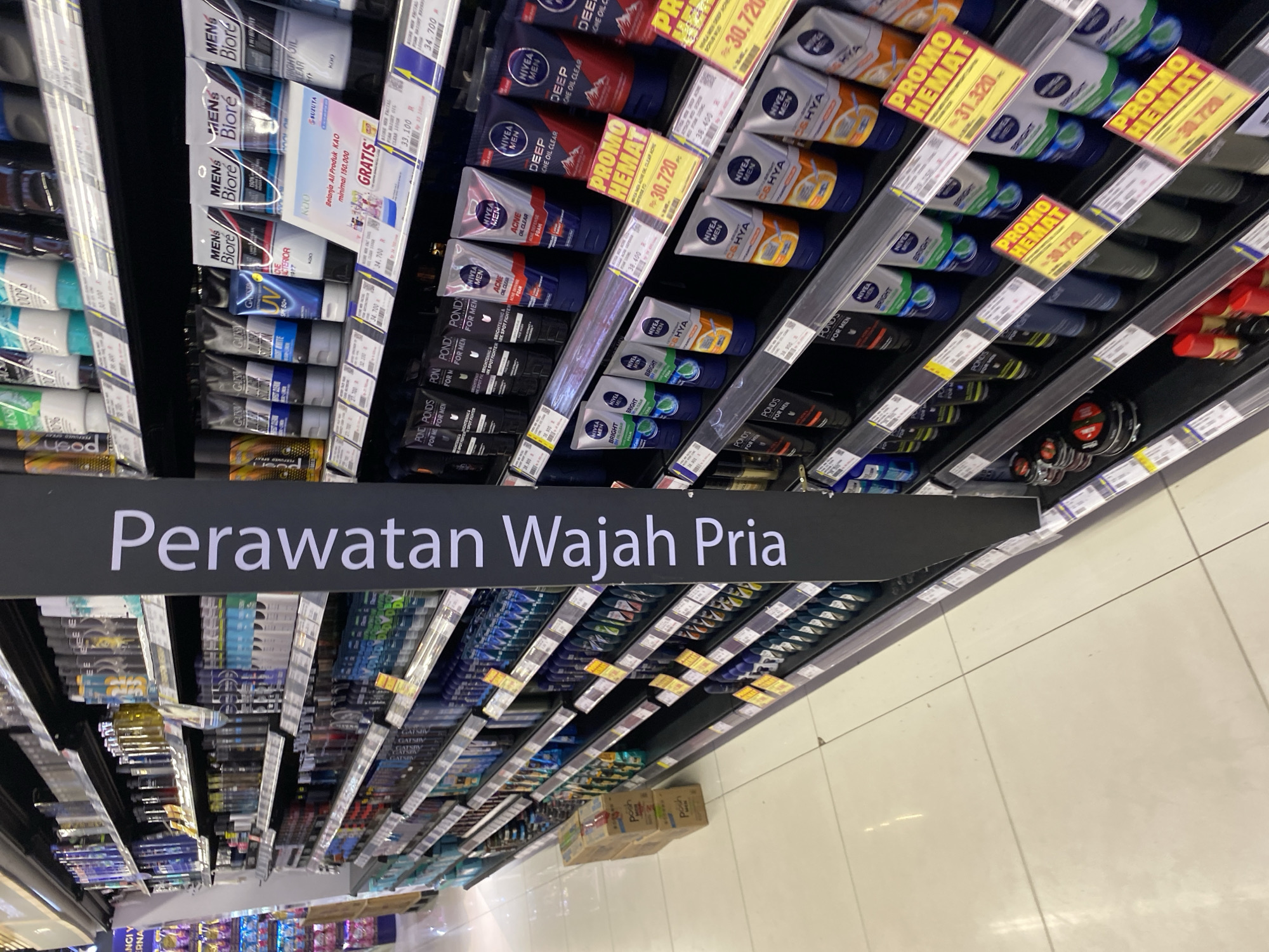 The image shows a well-lit supermarket aisle displaying men's skincare products. The shelves are organized with various brands and types of facial cleansers, toners, and other personal care items. A prominent black sign with white text in Indonesian reads "Perawatan Wajah Pria," which translates to "Men's Facial Care." Several products feature promotional tags, indicating discounts and special offers. The floor is tiled and clean. The general environment suggests a retail setting, likely a large supermarket or department store. The presence of "Banda Aceh, Indonesia" in the prompt indicates that this scene is located within that city.