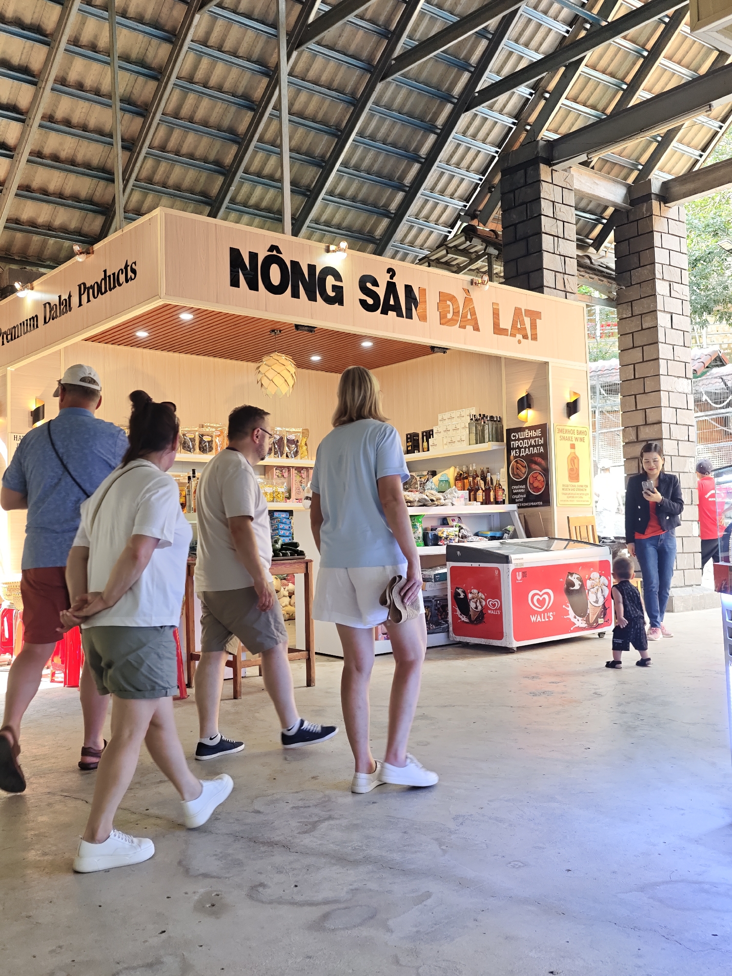 A group of people are gathered in an open-air market stall area. The stall is branded with "Premium Dalat Products" and "NÔNG SẢN ĐÀ LẠT". Inside the stall, shelves display various products, including bottles of liquid and packaged goods. An ice cream freezer with "Wall's" branding is visible in the foreground. The ground is paved with concrete. Several individuals are walking or standing around the stall. One woman in the background is looking at her phone. A young child is also present, standing near the ice cream freezer. The overall environment appears to be a commercial space, likely a tourist-oriented market. The location is identified as Định An, Vietnam.