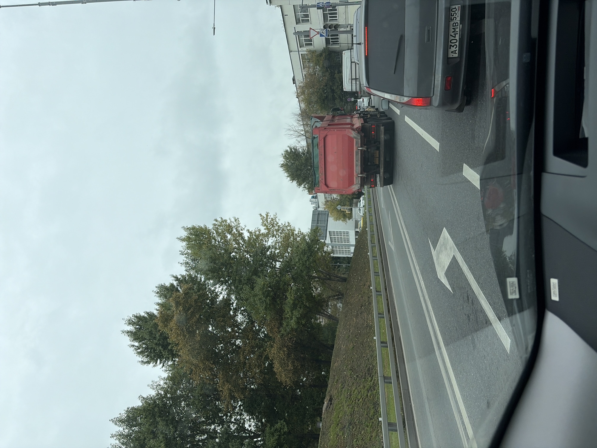 The image depicts a view from inside a vehicle traveling on a multi-lane road in Moscow, Russia, under an overcast sky. The road has clear lane markings, including a prominent left-turn arrow. Several vehicles are visible in front, including a red dump truck, a white truck, and a dark gray car. The gray car's license plate, which appears to be Russian, reads "A304MB 550". To the left of the road is a grassy embankment with trees, some of which are showing autumn foliage. In the background, buildings and traffic lights are visible. The scene suggests a typical urban traffic environment.