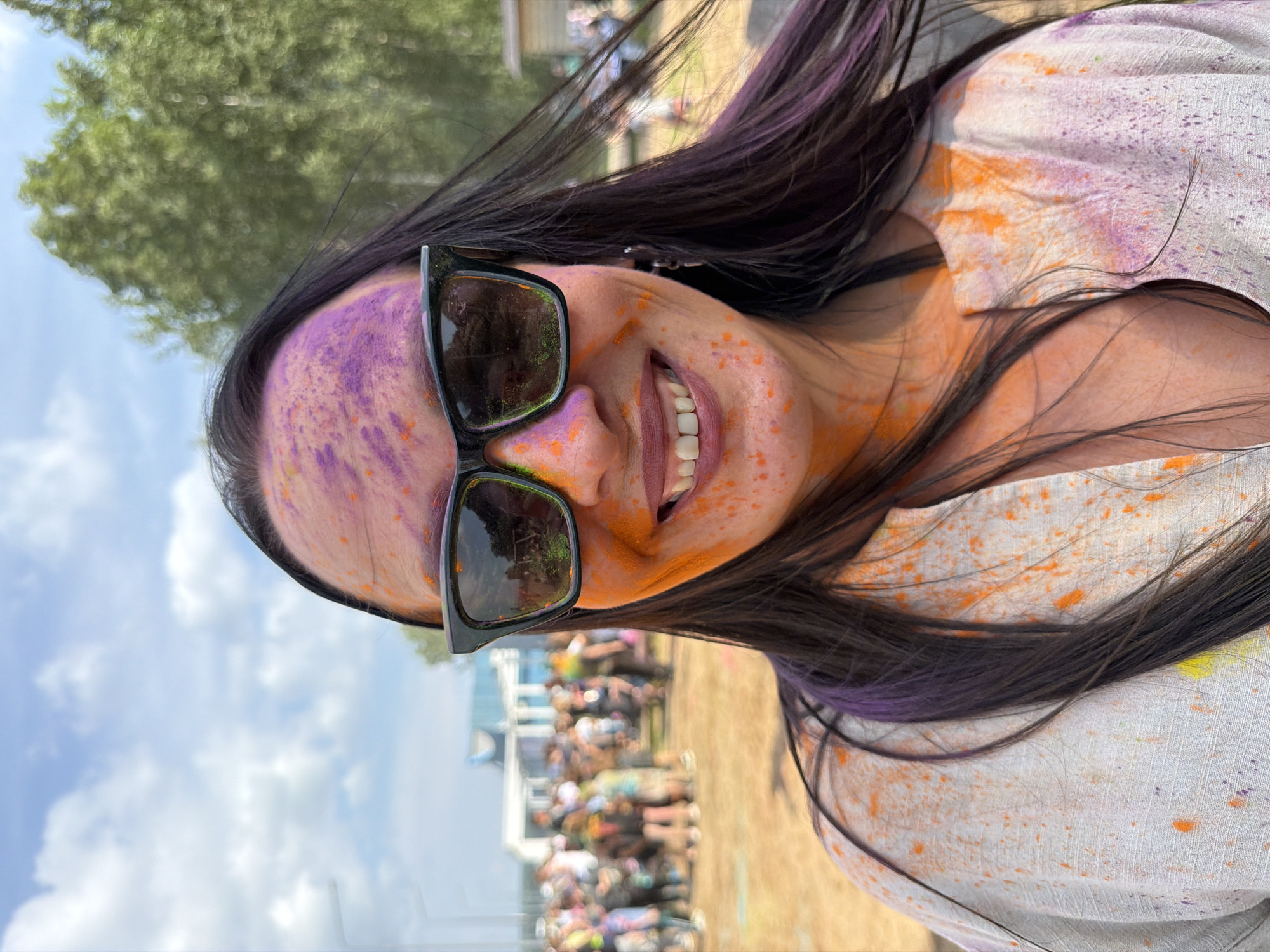 A woman, her face and upper body covered in streaks of orange and purple powder, smiles while wearing black sunglasses. She has long, dark hair with streaks of purple. She is outdoors in a public gathering, with a crowd of people visible in the background. Trees and a blue sky with clouds form the backdrop. The setting appears to be a festival or event, possibly the Holi festival, given the colored powder. This scene is likely taking place in the city of Chaikovsky, Russia, as indicated by the provided context.