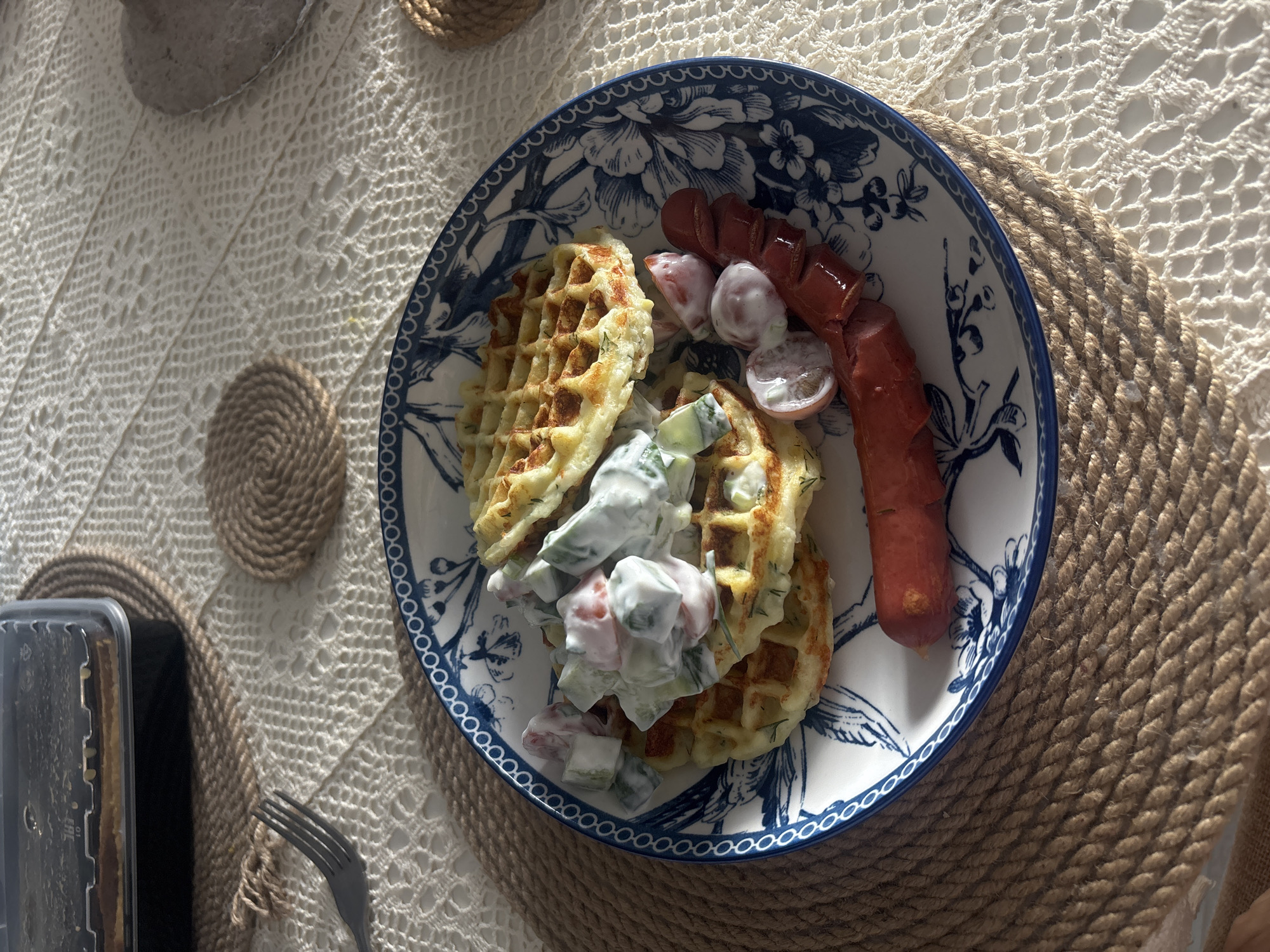 The image depicts a plate of food, containing waffles, a salad of cucumbers and tomatoes with a creamy dressing, and a sausage, on a table setting. The plate has a blue and white floral pattern. The table includes a crocheted white tablecloth, and a thick rope placemat. A fork is visible beside a dark, rectangular container. The image was likely taken indoors, in a home or restaurant setting. There is no evidence suggesting the location is in Krasnoyarsk, Russia.