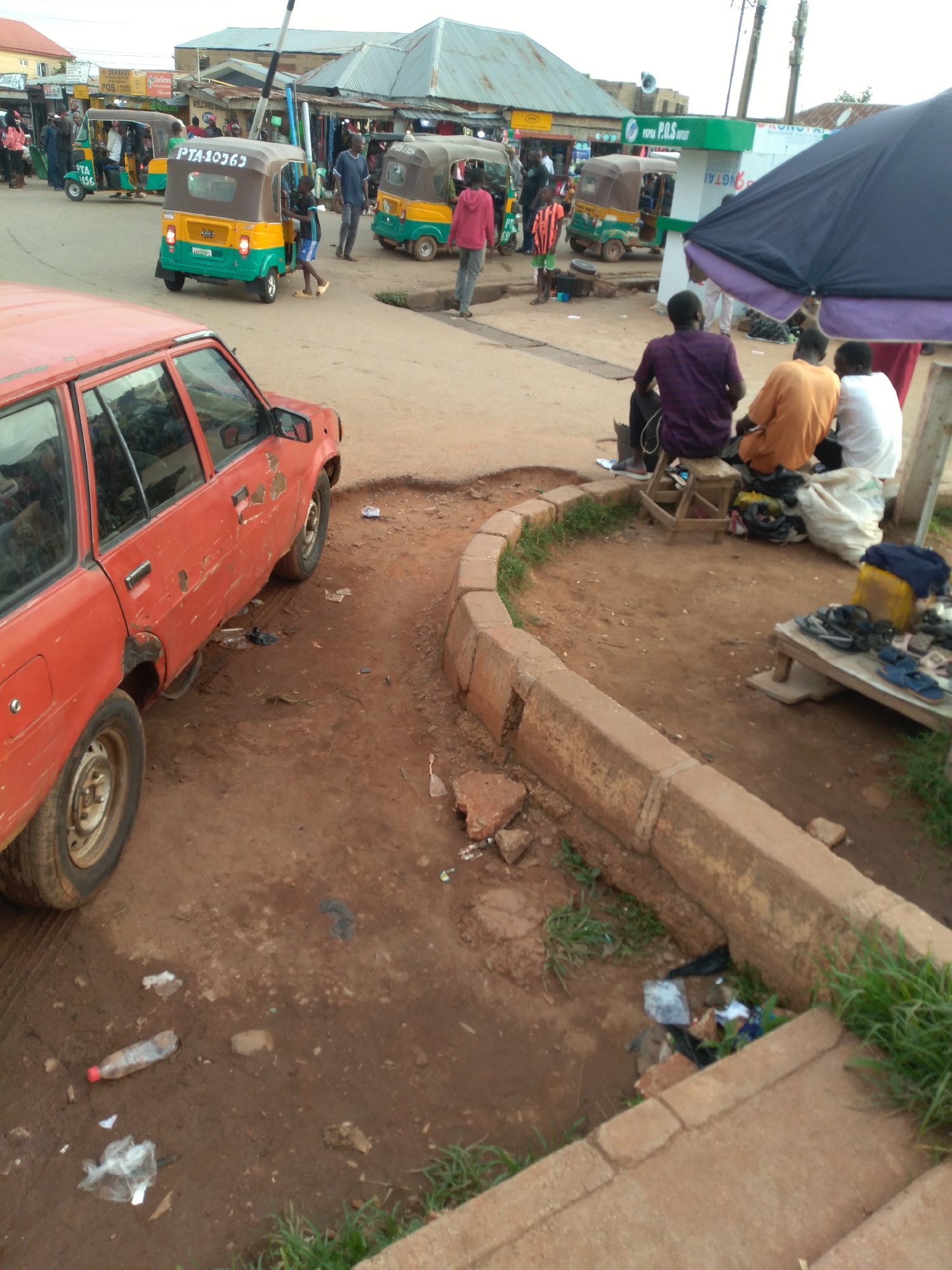 The image depicts a busy street scene in Gura Top, Nigeria, characterized by unpaved ground, a red station wagon, and several auto-rickshaws, locally known as "keke napeps". The auto-rickshaws, with their distinctive yellow and green colors, are a prominent feature, some appearing to be in motion or parked, suggesting a transport hub or busy thoroughfare. One keke napep displays the license plate "PTA 10363".
Several individuals are present, some appearing to be passengers or drivers, while others are seated by the roadside, possibly waiting or socializing. The setting suggests a commercial area with shops and stalls visible in the background, indicated by the presence of awnings and market-like structures. The ground is littered with debris, including plastic bottles and bags, highlighting a common environmental issue in urban areas. The overall atmosphere is one of active local life and commerce.