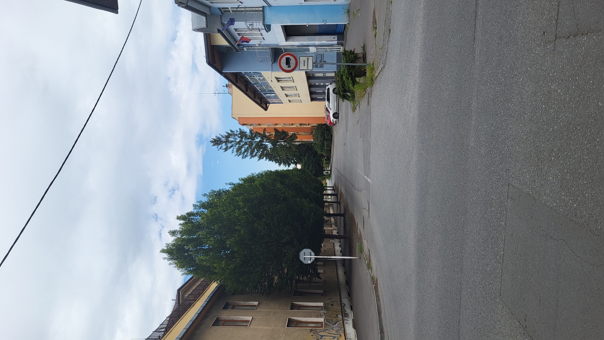 The image depicts a street scene in Prešov, Slovakia. On the left, a building with a yellow facade and windows is visible, exhibiting graffiti on its lower portion. A row of tall, mature trees lines the sidewalk in front of this building. The street itself is paved, with a visible white line marking a lane. On the right side of the street, a multi-story building painted in shades of blue and white stands, with a balcony and flags. A traffic sign indicating a restriction for trucks is prominently displayed. Adjacent to this building, parked cars, including a white SUV and a red car, are visible. Further down the street, other buildings in orange and yellow hues can be seen, along with more trees, including a tall conifer. The sky is partly cloudy.