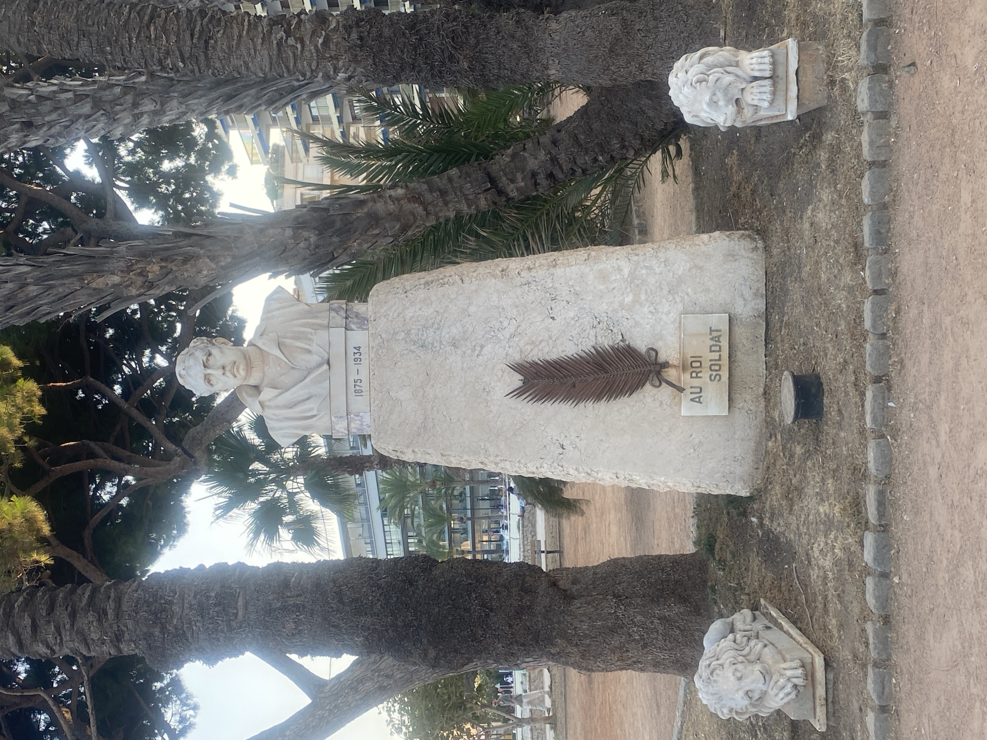 A bust of a man in civilian attire stands atop a stone monument in Antibes, France. The monument is flanked by two stone lions in a park setting with palm trees and pine trees. The bust appears to be made of marble and is mounted on a rectangular stone pedestal. A plaque below the bust reads "1875 - 1934." A metal palm frond is affixed to the front of the monument below the plaque, and another rectangular plaque reads "AU ROI SOLDAT." The overall scene depicts a memorial or statue in a public outdoor space.