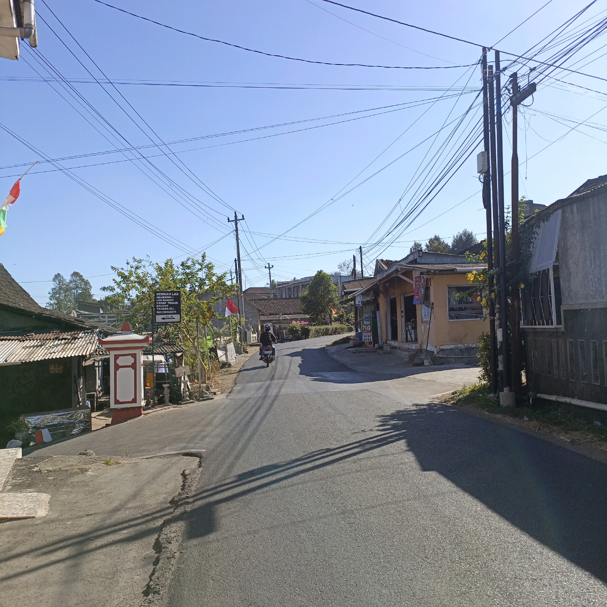 The image depicts a street scene in Candirejo, Indonesia, under a clear blue sky. A paved road runs through the center of the frame, with buildings and vegetation lining its sides. In the distance, several utility poles with a complex network of wires are visible.
On the left side of the road, a white and red ornamental pillar stands near a building with a corrugated metal roof. A signpost with text is planted next to it. Further down the street, a motorcycle is being ridden by an individual wearing a dark jacket and helmet, heading away from the camera.
On the right side of the road, a small building with an orange facade and open doorways is situated. Another building with a grey facade and a partially visible roof is adjacent to it. Flags are hung on poles in front of some of the buildings. The scene is illuminated by bright sunlight, casting distinct shadows across the road and ground. The overall impression is of a quiet, rural or semi-rural street in Indonesia.