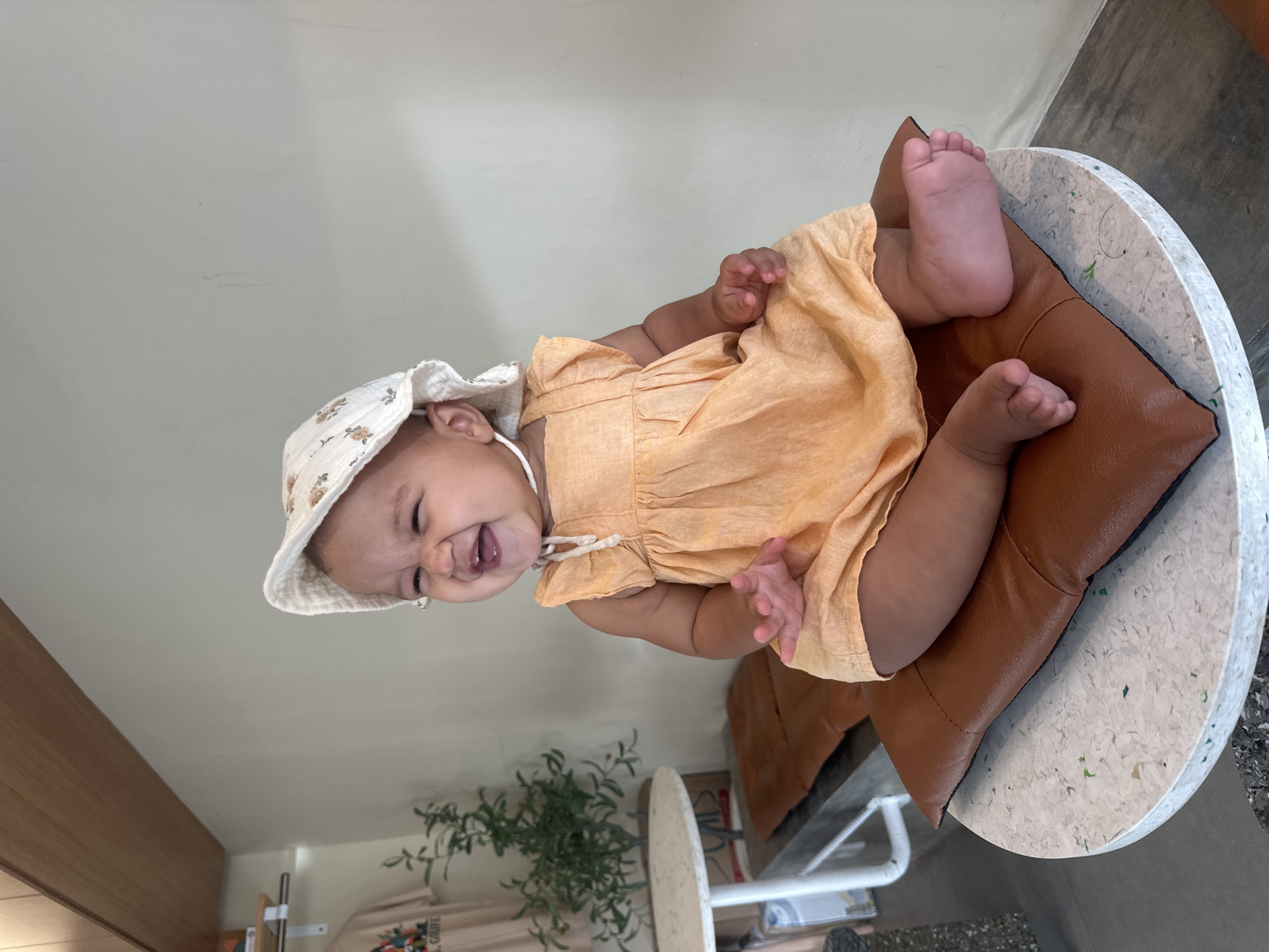 A baby wearing a floral bonnet and an orange romper is seated on a brown leather pillow, which itself rests on a round, mottled white stool. The baby is smiling and appears to be in a playful mood. The scene is indoors, likely in a building located in North Kuta, Indonesia. A small plant is visible in the background, along with a clothing rack. The lighting is bright and natural.