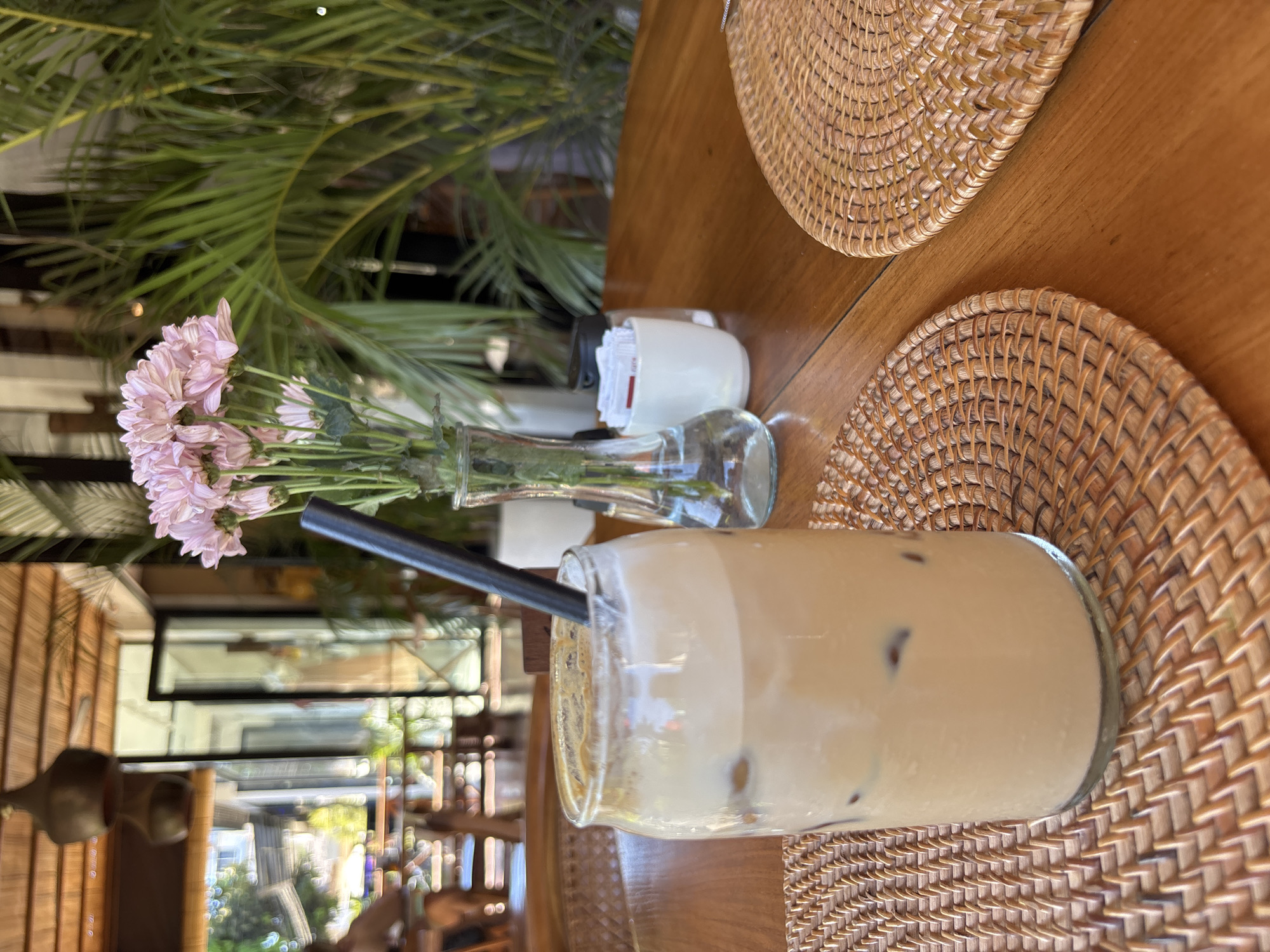 A table is presented indoors, near a window offering a view of the outdoors with trees visible. On the table, there's a glass of milky coffee with ice and a black straw, resting on a round, woven straw placemat. Nearby, a small vase holds pink flowers, accompanied by a container of sugar packets. Another similar placemat is also on the table. The setting appears to be in Pecatu, Indonesia, as indicated by the geotag.
