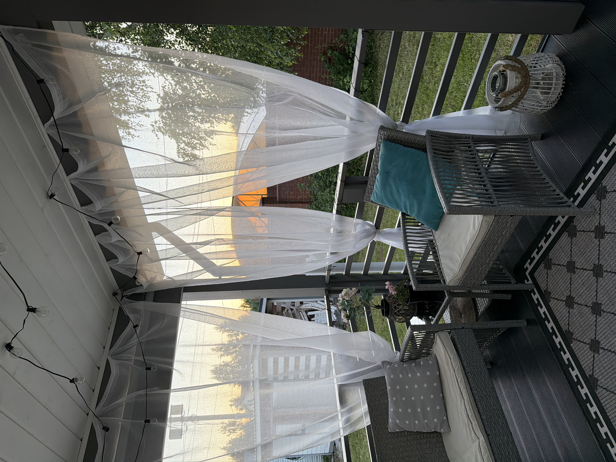 The image depicts a covered patio or porch in the city of Alyoshkino DP, Russia, during the evening golden hour. The setting appears to be a residential area, possibly a dacha (summer house) given the outdoor furniture and rustic elements.
The foreground is dominated by a dark wooden deck, partially covered by a patterned rug. On the deck are two wicker chairs. The chair on the left has a grey cushion with white stars and a light grey seat cushion. The chair on the right has a turquoise cushion and a light grey seat cushion. Between the chairs is a small dark table holding a dark vase with pink flowers. To the right of the right chair, a white woven lantern with a rope handle sits on the deck.
Behind the furniture, a metal railing encloses the porch. Beyond the railing, a grassy area is visible. The porch ceiling is white wood, from which string lights with exposed bulbs are hung. Translucent white curtains are draped from the ceiling, obscuring the view of the surroundings but allowing the warm glow of the sunset to filter through. In the background, beyond the curtains and railing, there are glimpses of buildings and trees silhouetted against the bright orange and yellow sky. The overall atmosphere is peaceful and inviting, suggesting a relaxed outdoor living space. There are no people present in the image.