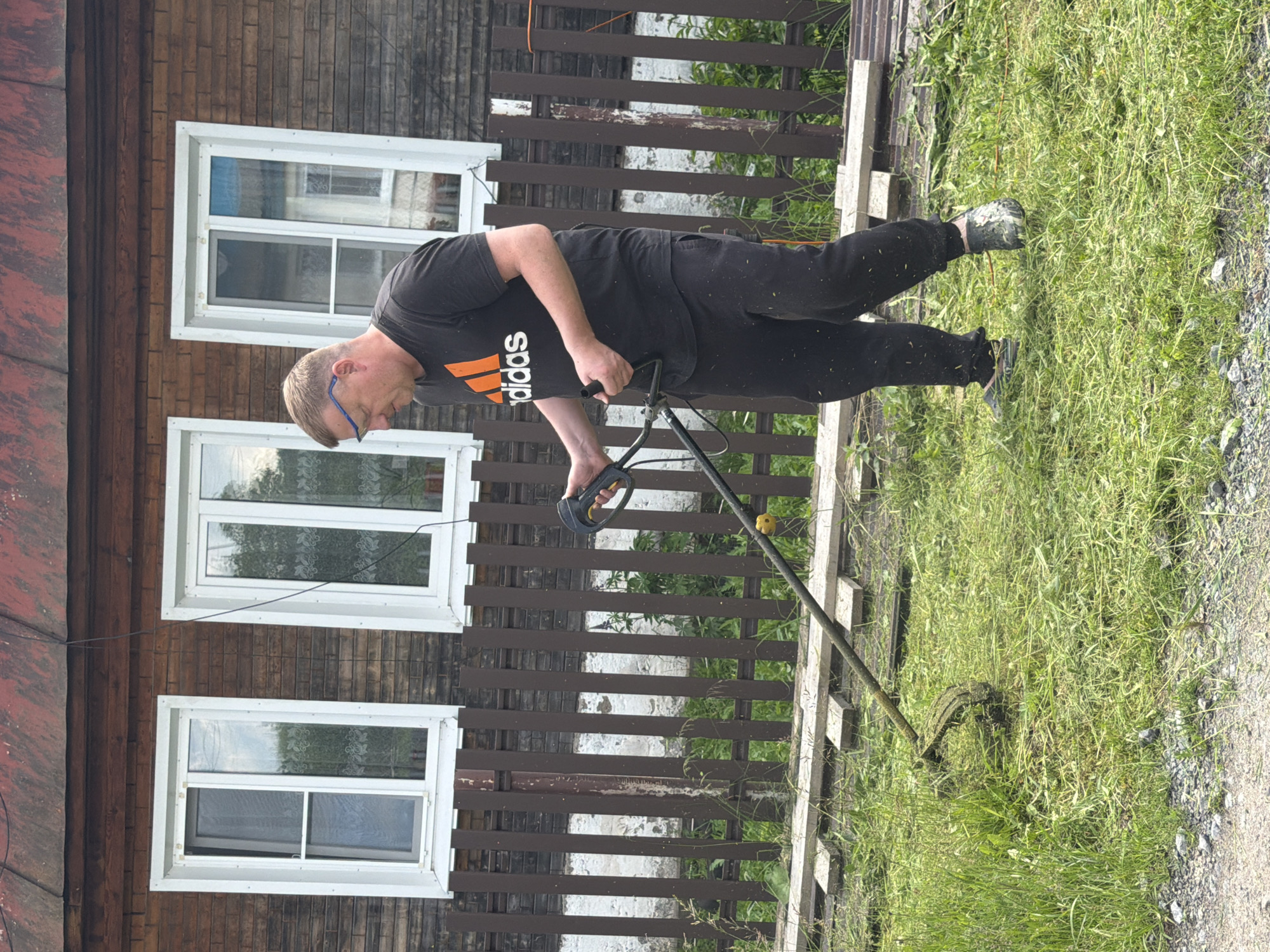 A man is mowing a lawn in Mezhevoy, Russia. The man, wearing a black Adidas t-shirt and black sweatpants, is using a string trimmer to cut tall grass in front of a rustic wooden house. The house has three windows and a brown wooden fence runs along the property. The ground is covered in cut grass and some gravel.