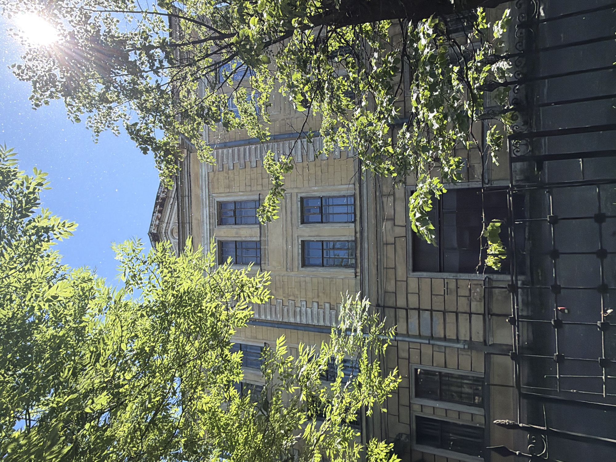 This image captures a sun-drenched view of the facade of an old building in Saint Petersburg, Russia, framed by lush green trees. The building itself is a light yellow color, adorned with classical architectural elements such as pilasters and decorative moldings. Several windows, some with reflective blue glass, are visible on its multiple stories. A dark, large garage-like door occupies a significant portion of the lower level. In the foreground, a black metal fence with ornate scrollwork runs across the bottom of the frame. The sky above is a vibrant blue, with the sun casting strong rays and lens flares through the leaves of the trees, suggesting a bright, sunny day. The overall impression is one of urban architecture softened by abundant natural foliage and bright sunlight.