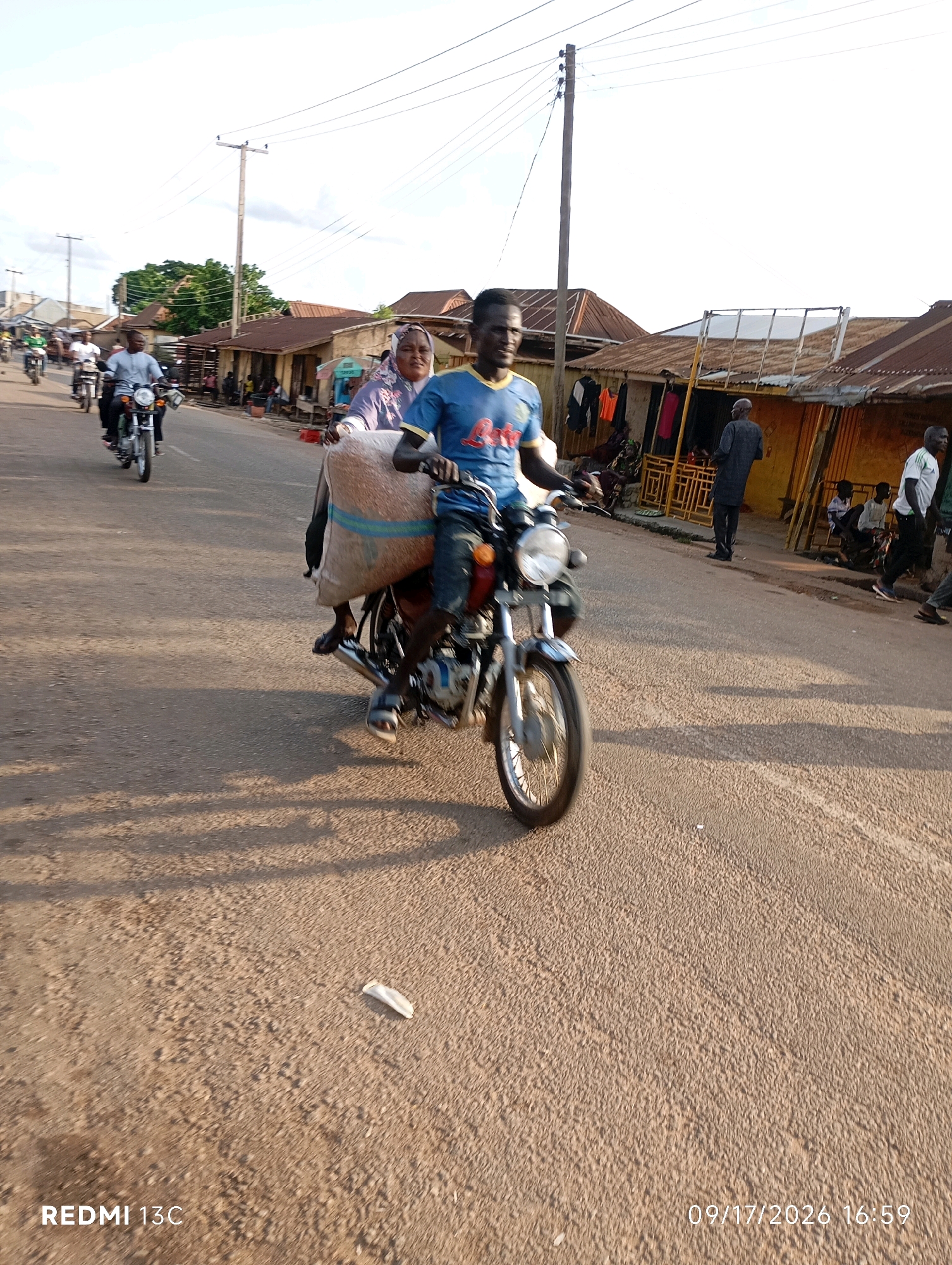In Shendam, Nigeria, a man in a blue t-shirt rides a motorcycle carrying a large sack of goods on the back. Beside him, a woman wearing a headscarf is seated on the motorcycle. The motorcycle is moving down a paved street lined with market stalls. The sky above is overcast, and shadows of people and objects are cast on the road. Other motorcycles and pedestrians are visible in the background, indicating a busy street scene. The timestamp 09/17/2026 16:59 is displayed in the lower right corner.