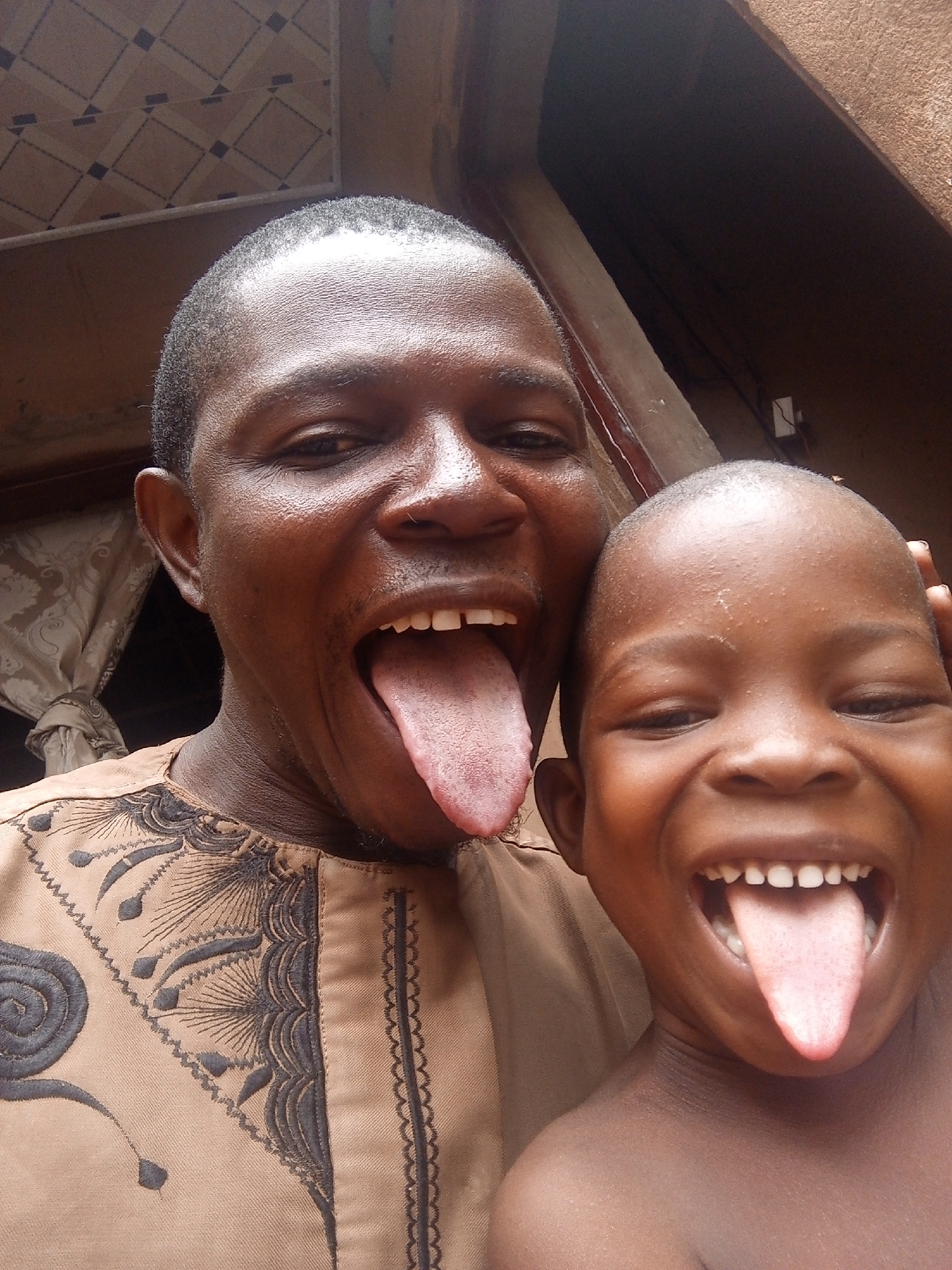 Two dark-skinned individuals, an adult and a child, are posing for a selfie outdoors in Tempure, Nigeria. Both have their mouths wide open, tongues extended, and are smiling broadly. The adult is wearing a light brown, embroidered garment. The child appears shirtless. The background includes patterned ceiling tiles, a curtain, and a partially visible building exterior.
