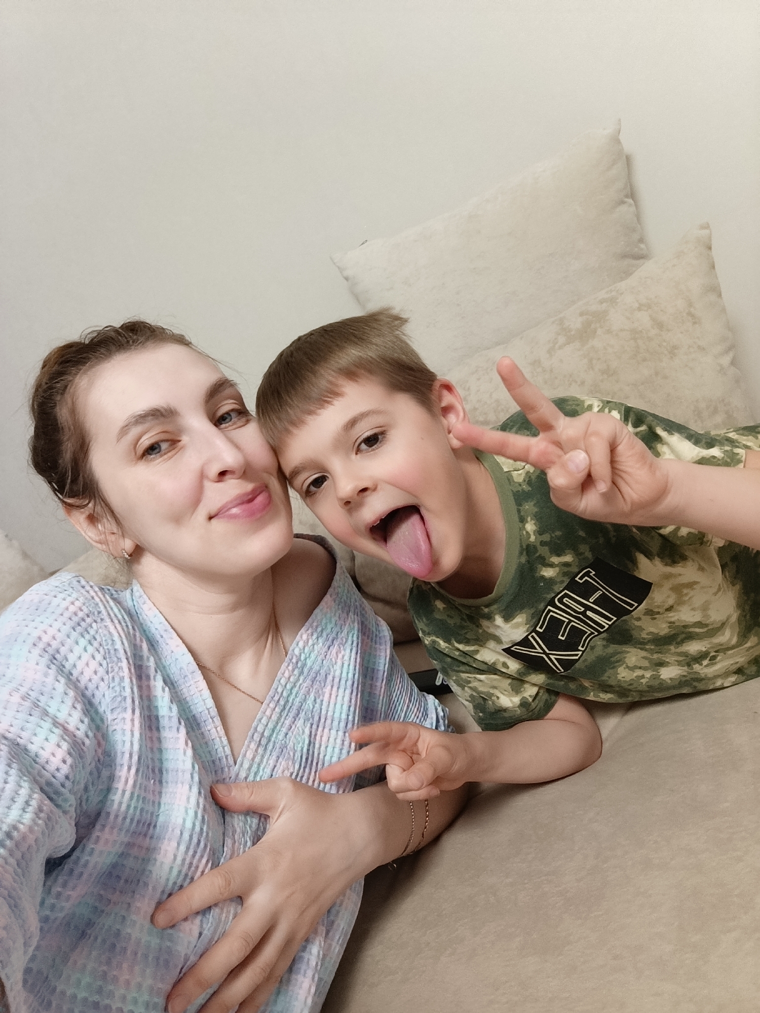 The image shows a selfie taken indoors, apparently in a private residence. A woman with light skin, brown hair pulled back, and wearing a pastel-colored robe is positioned on the left side of the frame. To her right is a young boy with short, light brown hair, wearing a camouflage patterned shirt. The boy is sticking his tongue out and making a "peace" sign with his right hand. The woman is also making a "peace" sign with her left hand. Both are smiling or grinning at the camera. The background features light-colored cushions and a wall. The subjects are positioned on a light brown sofa. There is no verifiable evidence to suggest that the location is in the city of Chaikovsky, Russia.