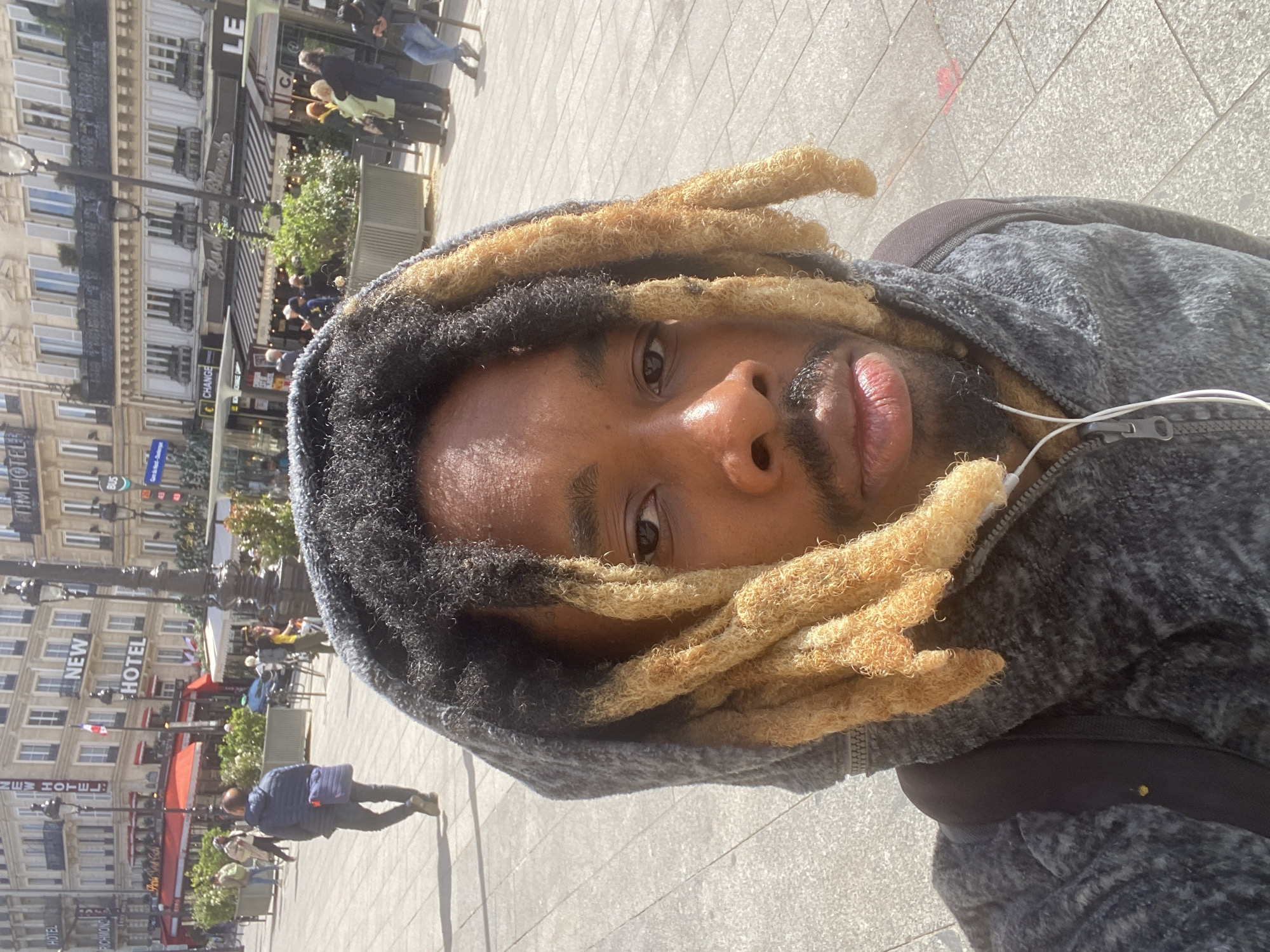 A close-up selfie features a person with dark skin, wearing a grey textured hooded jacket and white wired earphones. Their hair is styled in dreadlocks, black at the roots and blonde for the majority of their length. The person looks directly at the camera with a neutral expression.
The background shows a bright, sunny urban street, characteristic of Paris, France. Multi-story buildings with classic European architecture, featuring windows, shutters, and balconies, line the street. Several establishments have striped awnings, and people are visible walking on the paved ground and sitting at outdoor cafes.
The strong sunlight indicates a clear day, likely mid-morning or afternoon. Visible text includes "HOTEL" and "NEW HOTEL" on buildings, "LE" on another structure, and a "CHANGE" sign. An awning partially displays "Boulangerie La Maison Blanche." A blue bus stop sign with "BUS" is also discernible, and French flags are seen on some buildings. The image captures a lively street scene from the perspective of the main subject.