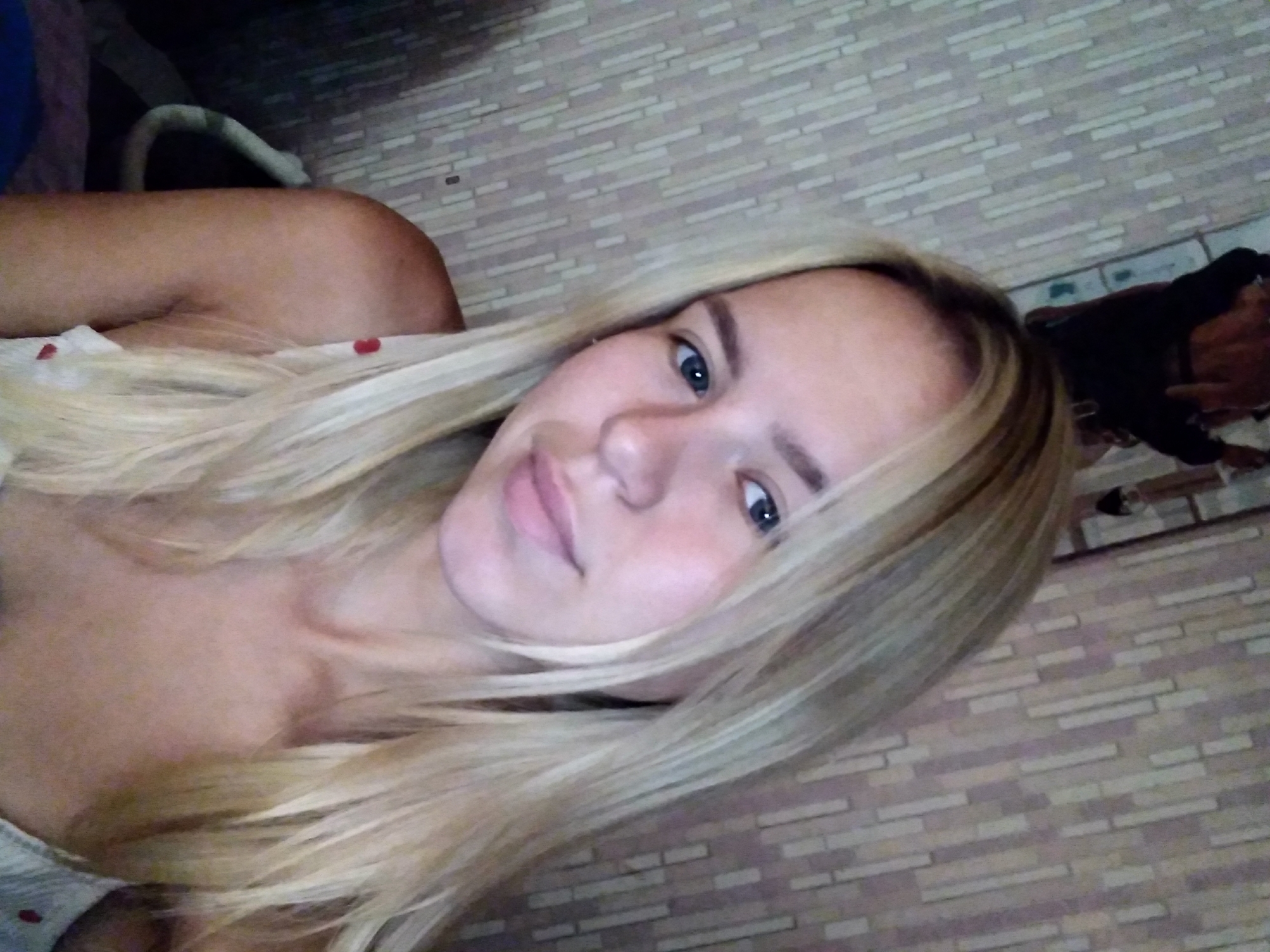 The image is a close-up selfie of a young woman with blonde hair, taken indoors. She is wearing a light-colored top with red polka dots. The background appears to be a wall with a patterned wallpaper. The overall lighting suggests it is likely daytime, but the room is not brightly lit. There are no specific indicators of a location within the city of Radisne, Ukraine.