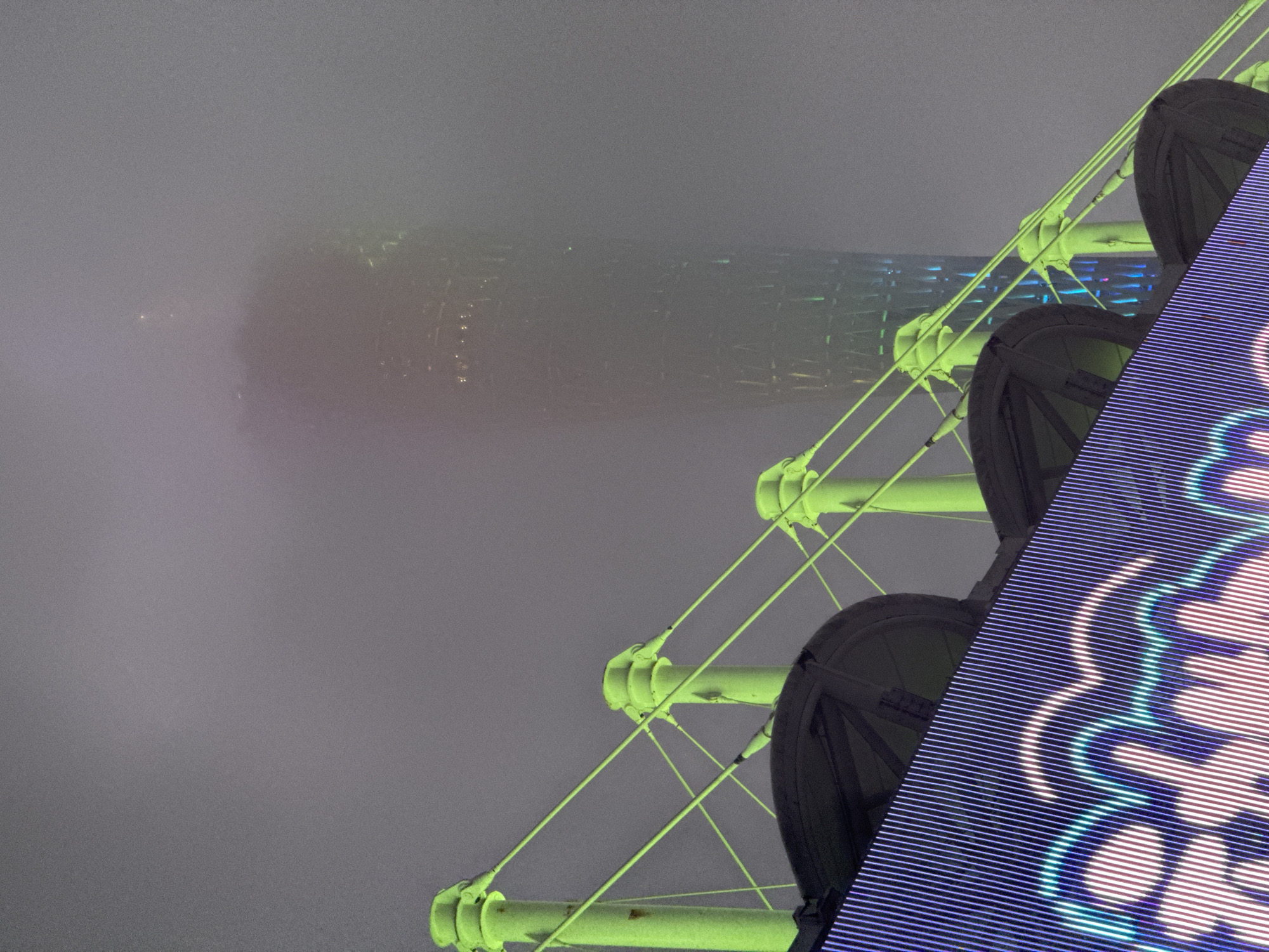 A nighttime, eye-level shot shows the Canton Tower obscured by fog, in the Tianhe District, China. The tower is faintly visible in the background, shrouded in a dense fog. In the foreground, a portion of a Ferris wheel structure is visible, with bright green support beams and visible black passenger cars. A digital screen with a lit design, in shades of blue and white, takes up a large portion of the right side of the frame. The scene is muted due to the foggy conditions.