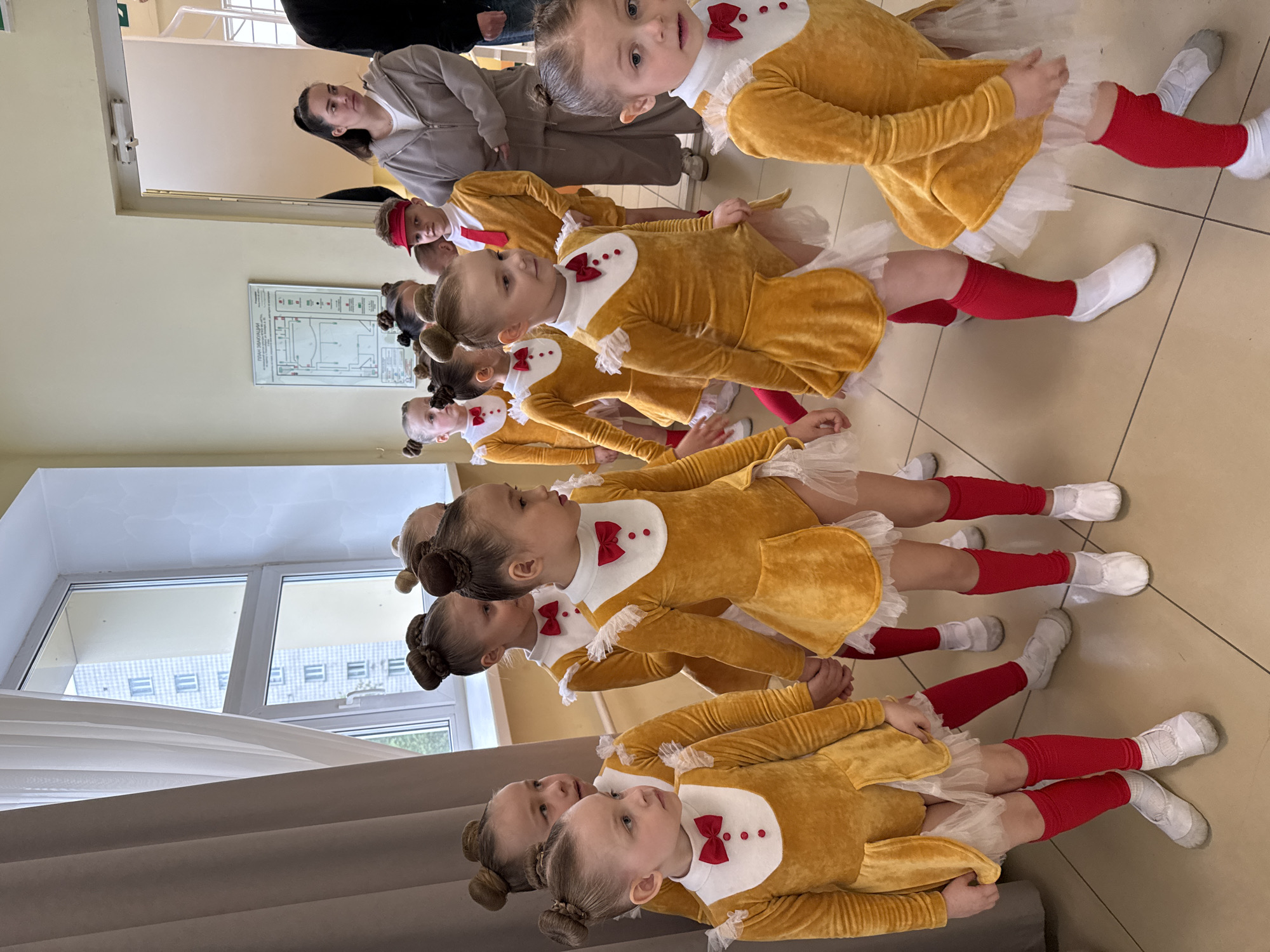 A group of young girls, likely ballet students, are lined up in a room. They are wearing identical yellow velvet dresses with white ruffled collars and red bow ties, red leg warmers, and white ballet shoes. Their hair is styled in buns. They are facing to the right. In the background, a woman in a grey jacket is observing them. A window and a door are visible in the background. Based on the presence of a sign in Russian ("План Путей эвакуации из здания"), it can be determined that this image was likely taken in Russia, and more specifically, it was taken in the city of Novosibirsk, Russia based on the building outside the window. The room appears to be a dance studio or a rehearsal room. The girls seem to be preparing for or recovering from a performance.